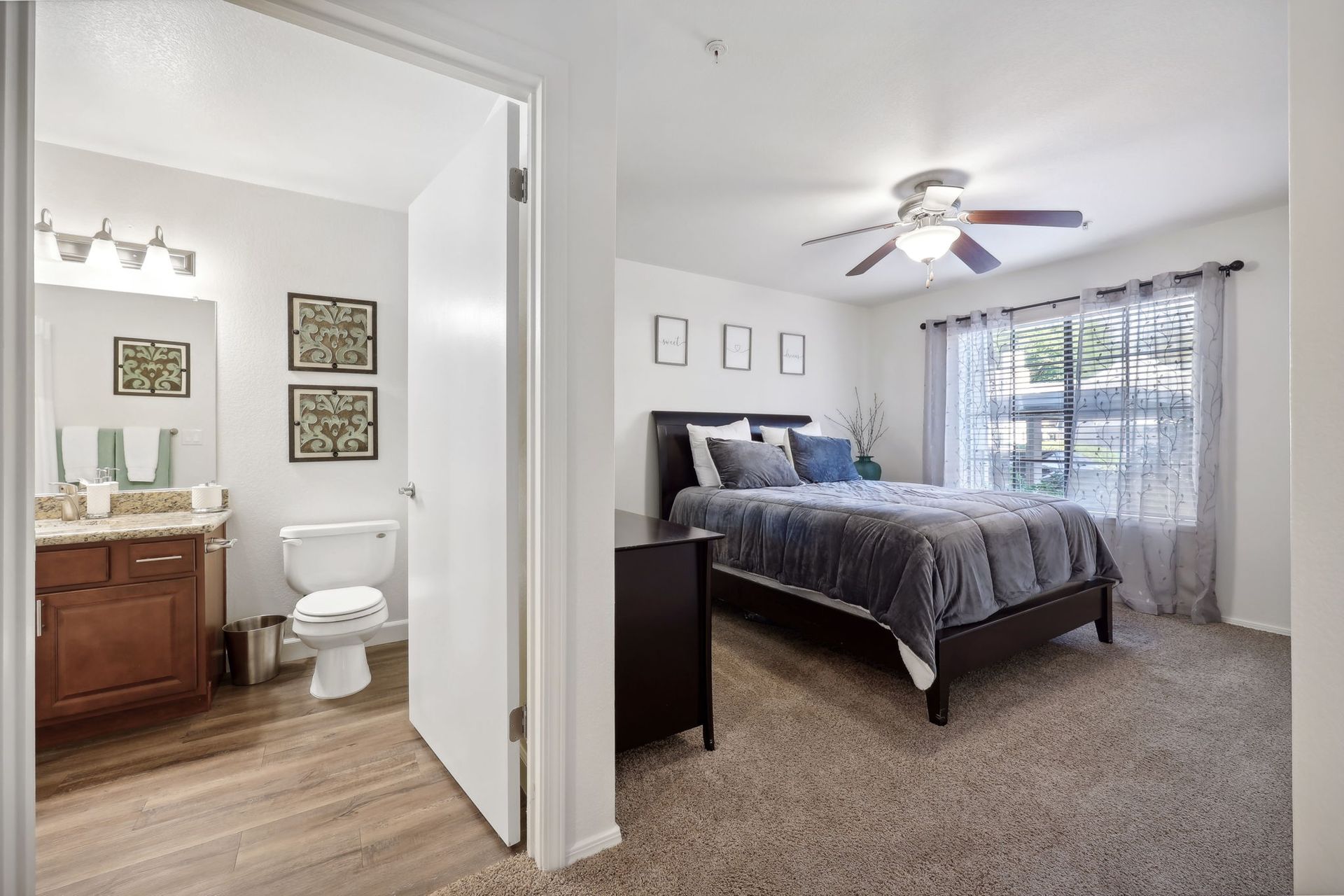 Bedroom with attached bathroom. Dark bed frame, gray bedding, brown carpet, window with sheer curtains.