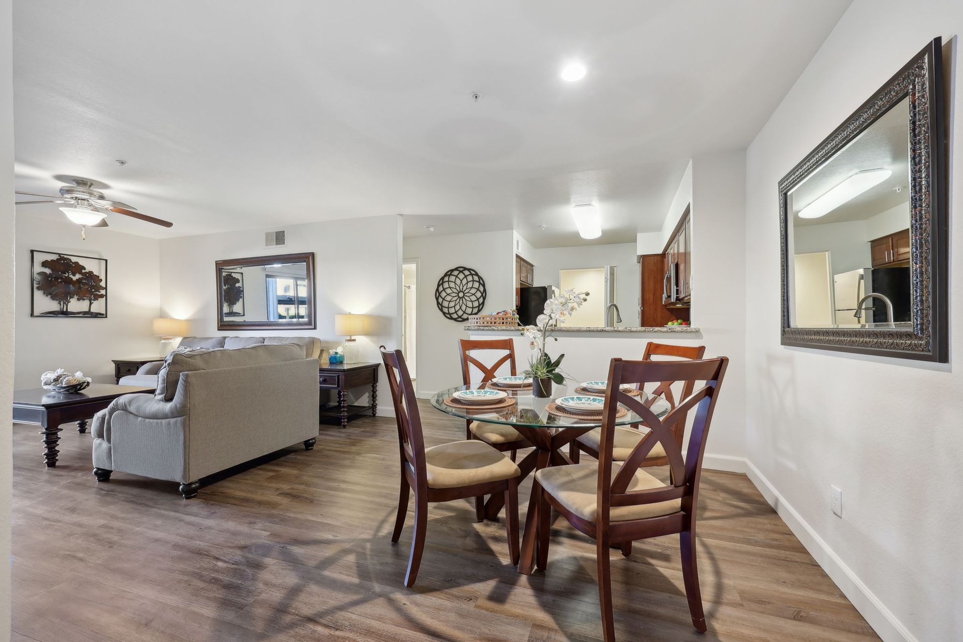 Dining area with glass table, four chairs, mirror, open to living room with sofa.