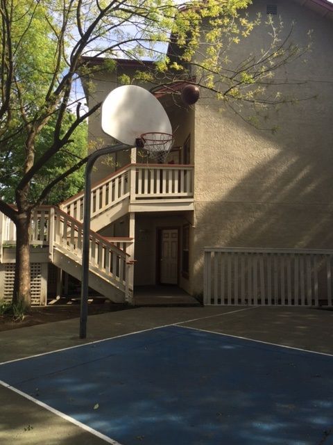 Basketball hoop on a blue court with a building in the background. Stairs lead to a doorway.