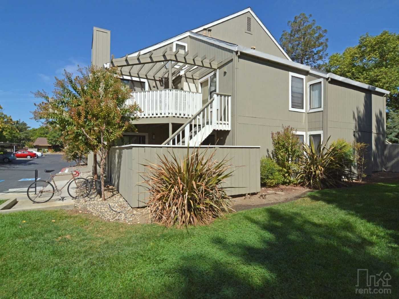 Two-story apartment building with stairs and a pergola; a bicycle sits near the building on a sunny day.