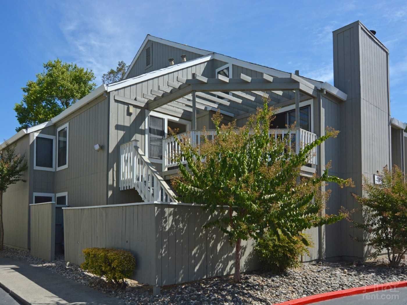 Gray two-story house with pergola, white stairs, and lush bushes under a blue sky.