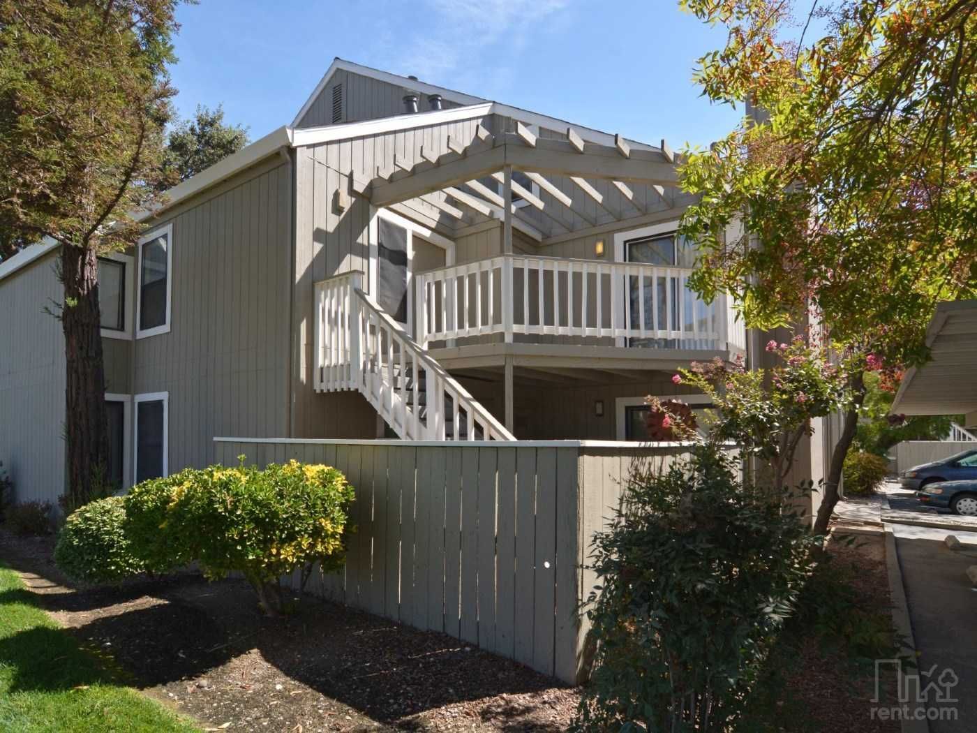 Two-story house with a wooden deck and pergola; grey exterior, surrounded by trees and a fence.