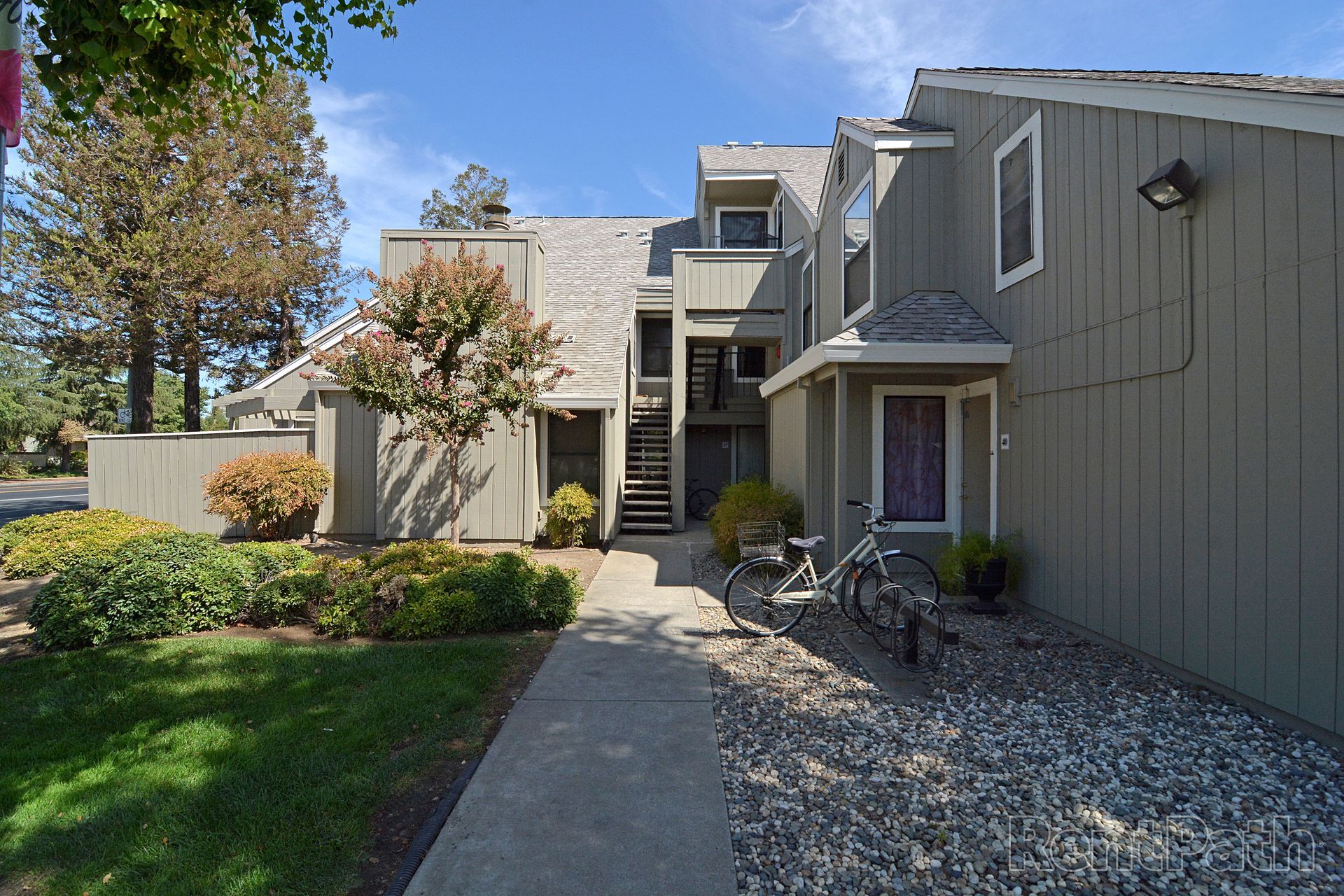 Apartment building exterior, gray siding, walkway, parked bicycle, landscaping with shrubs and trees.