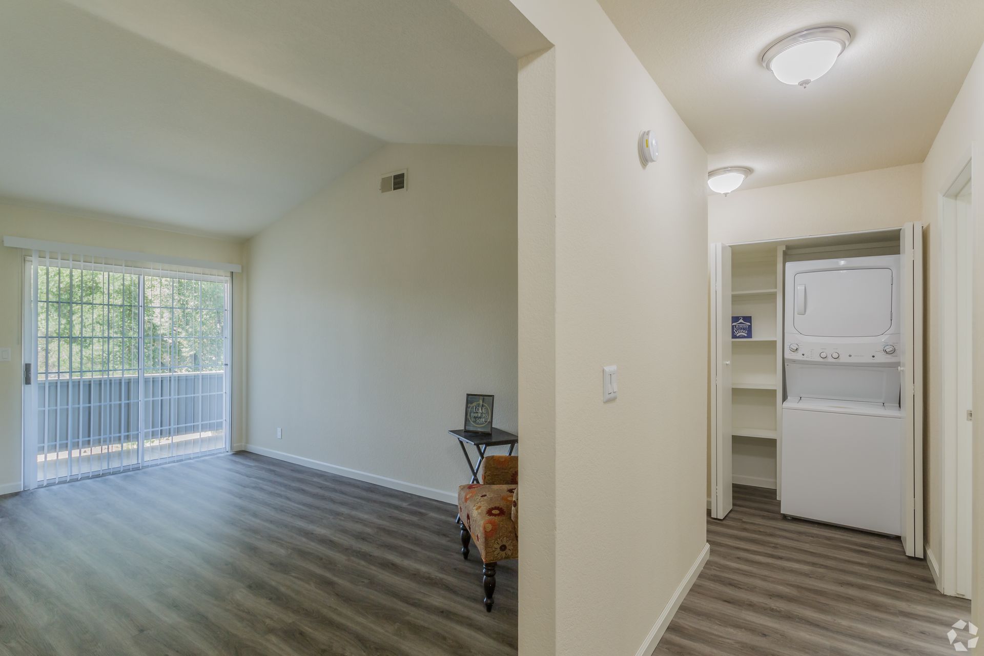 Interior view of an apartment: living room with balcony access, hallway with stacked washer/dryer, and neutral walls.