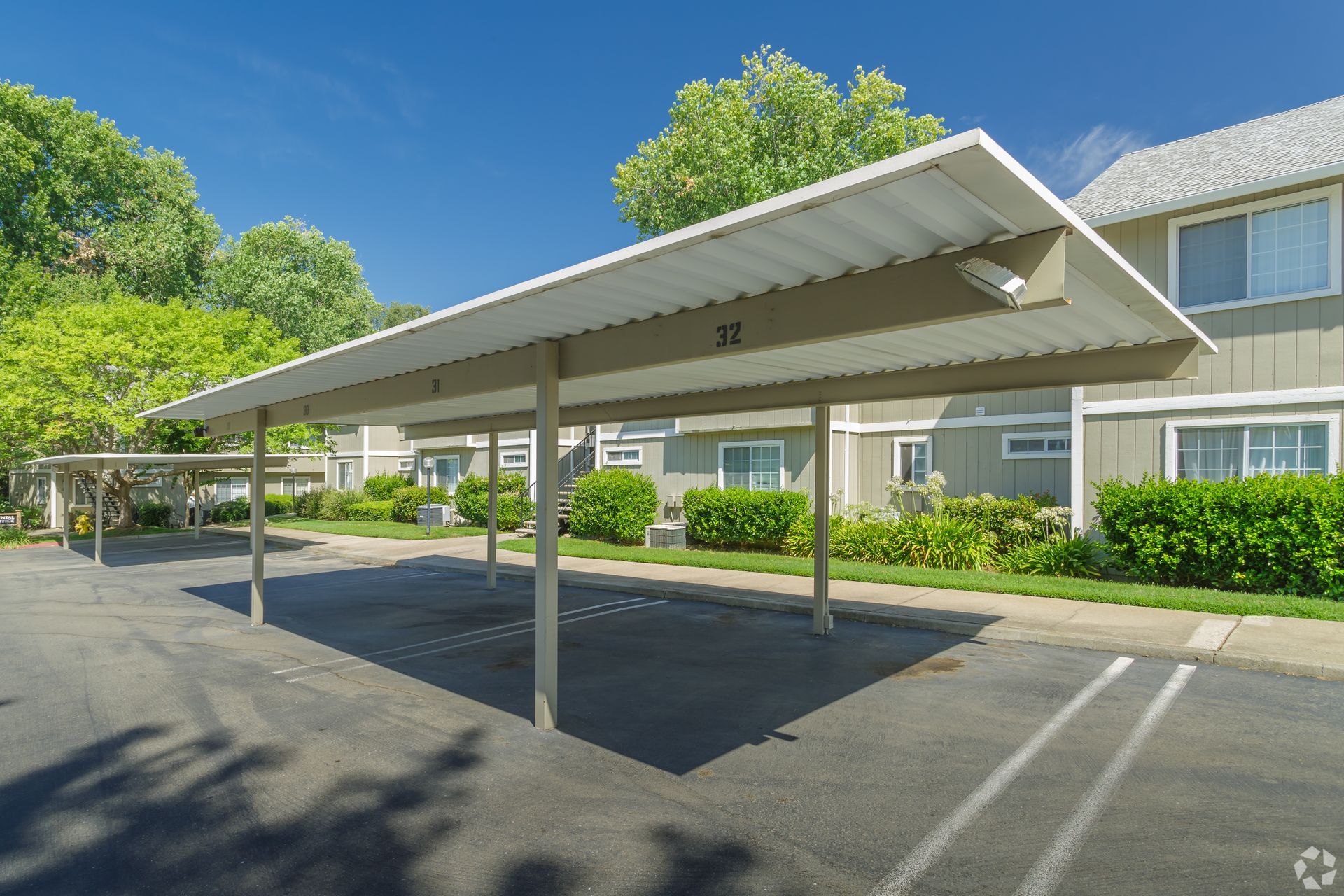 Carport with metal roof and support poles, next to apartment building and parking spaces.