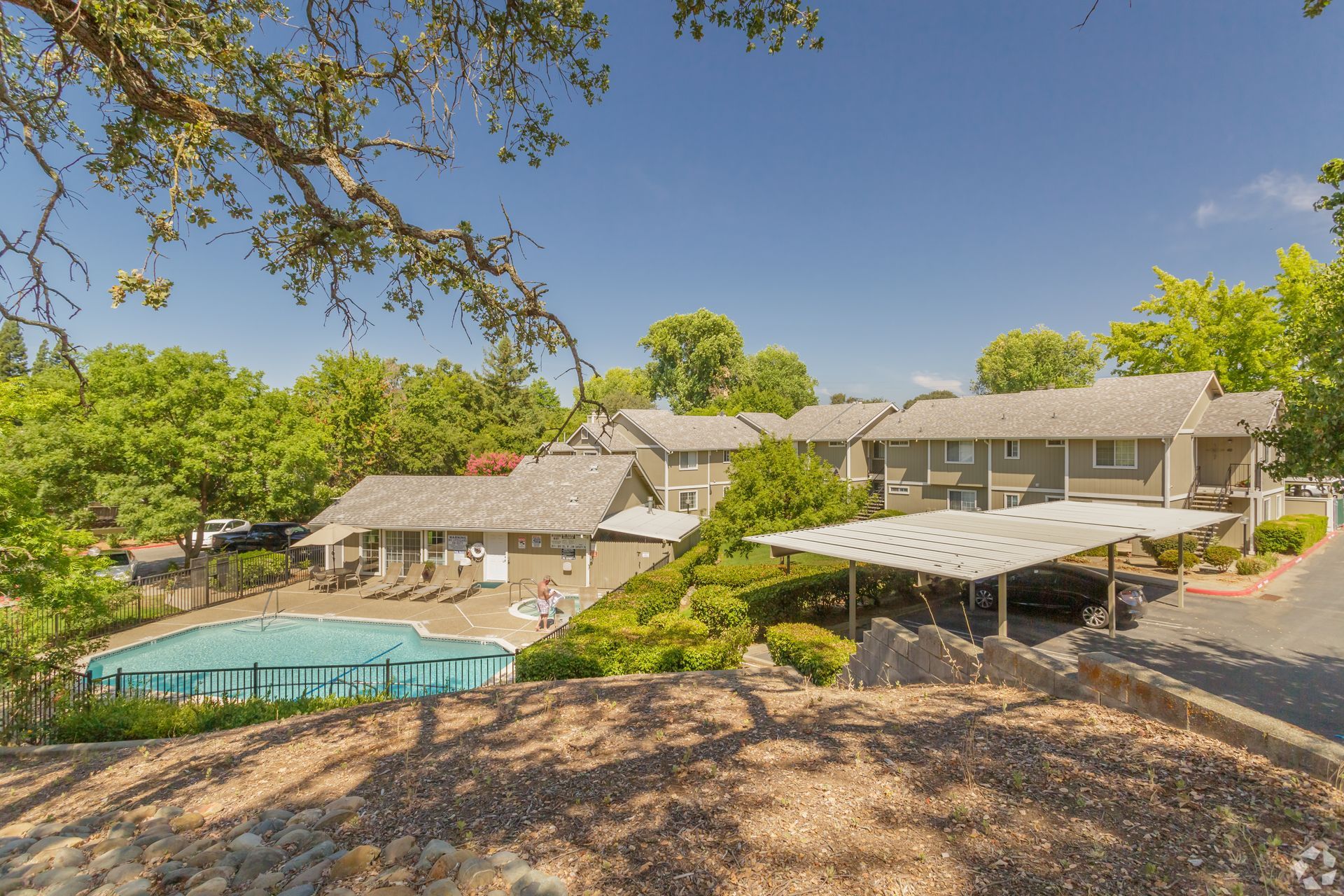 Apartment complex with a pool, carports, and lush greenery under a bright blue sky.