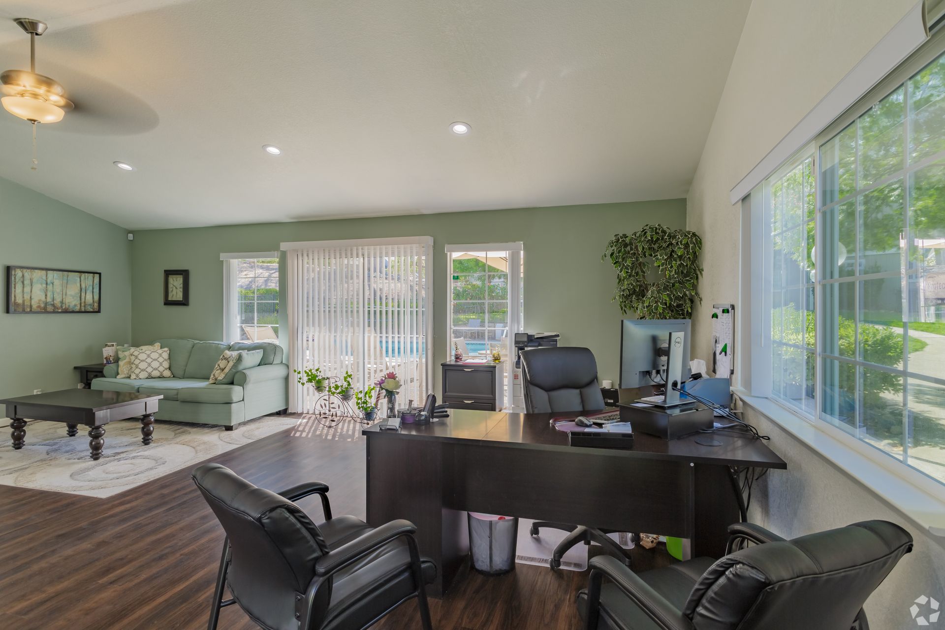 Office interior with desk, chairs, and couch. Windows offer a view of greenery.