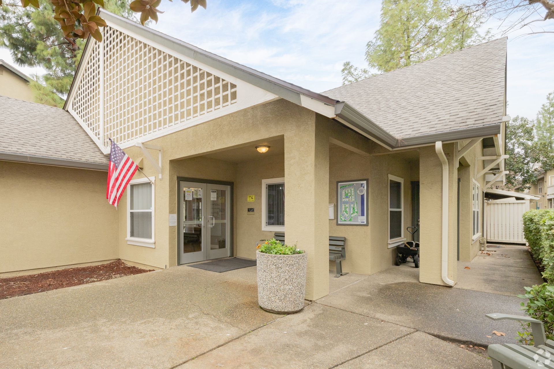 Tan building with angled roof, front entrance, US flag, concrete walkway, and decorative trellis.