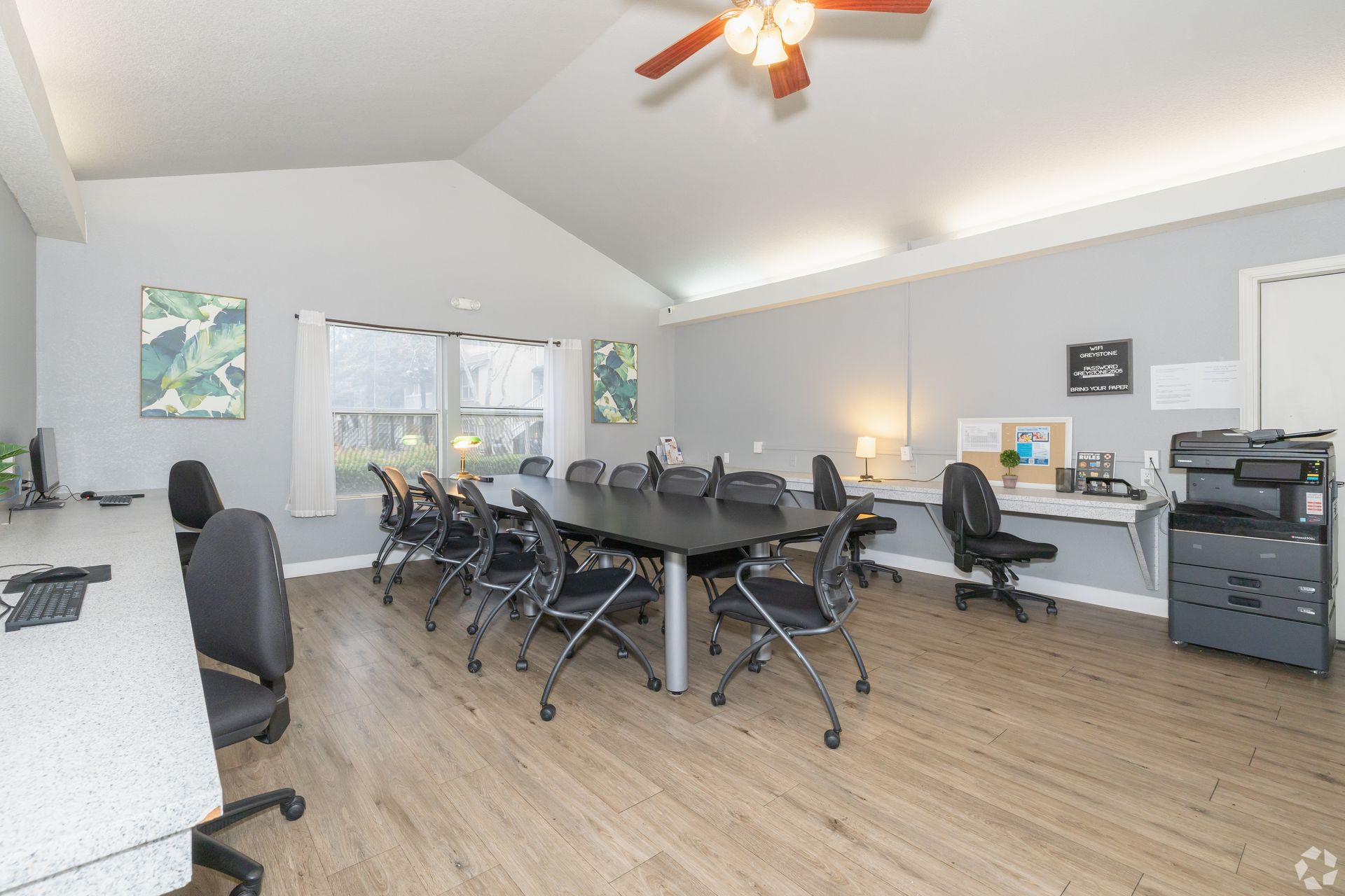 Conference room with long table, chairs, desks, printer, and ceiling fan. Light gray walls, wood floors.