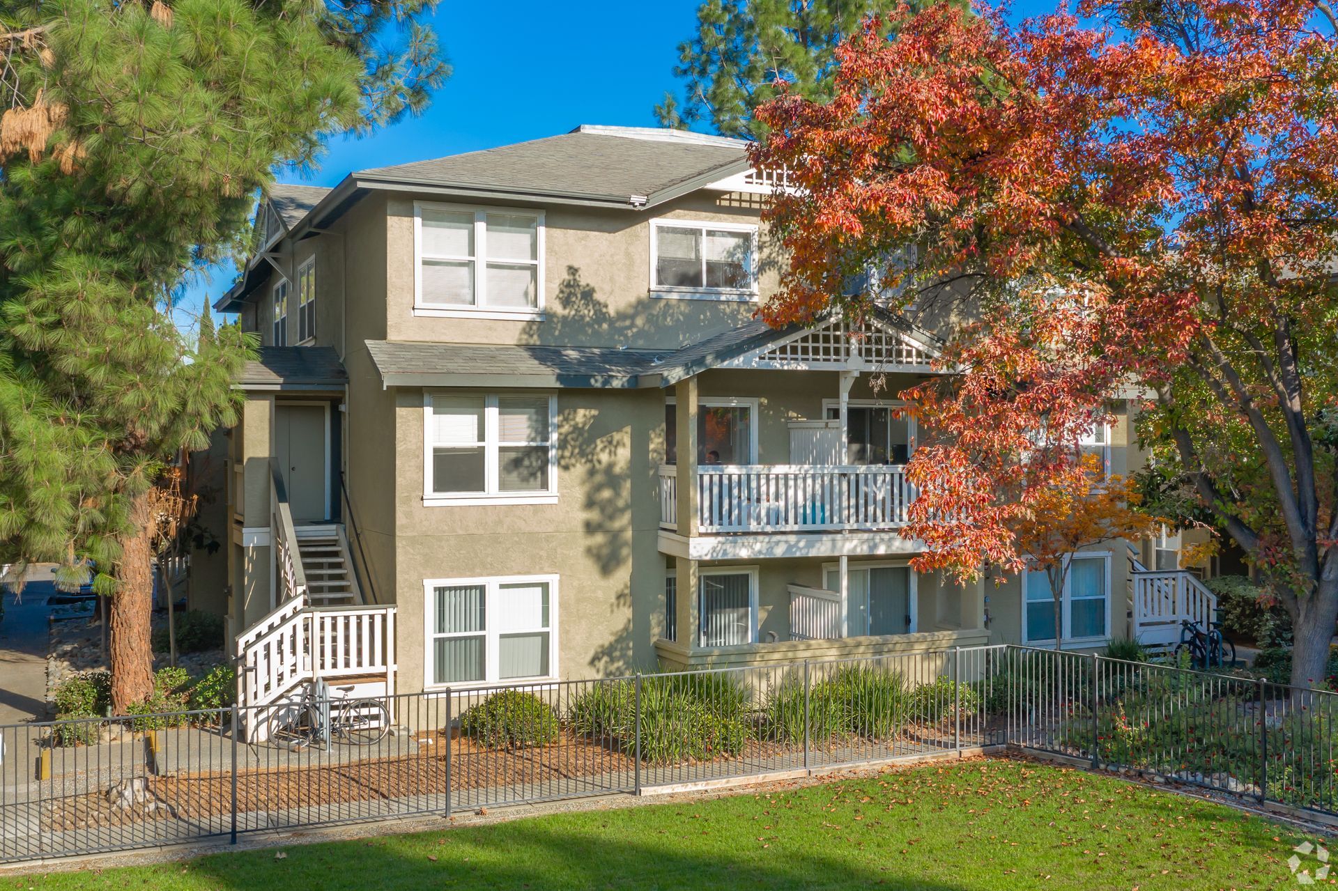 Three-story apartment building with balconies, beige exterior, surrounded by trees and a fenced lawn.