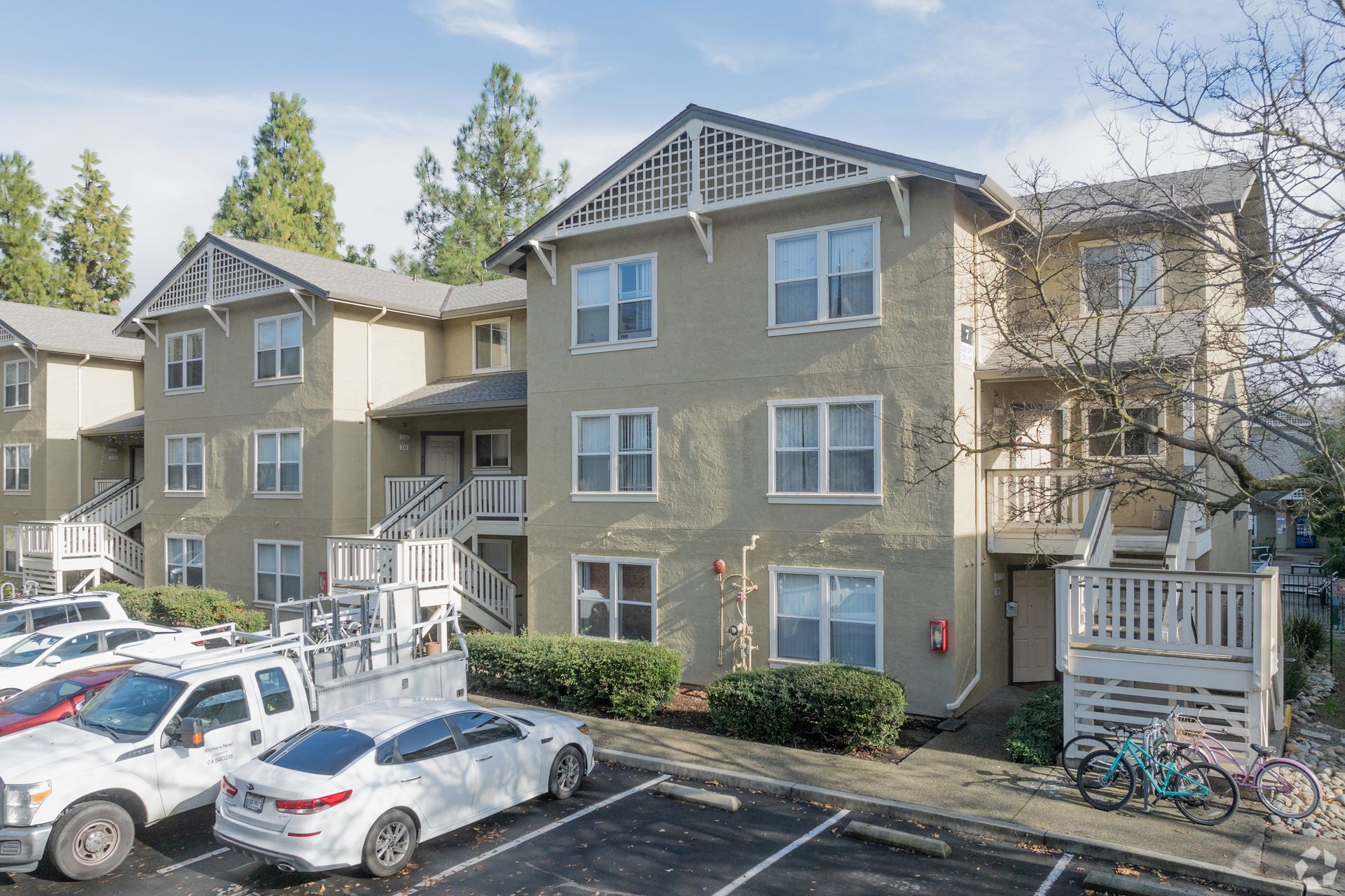 Apartment building exterior, three stories, beige stucco, parking lot with cars. Trees in the background.
