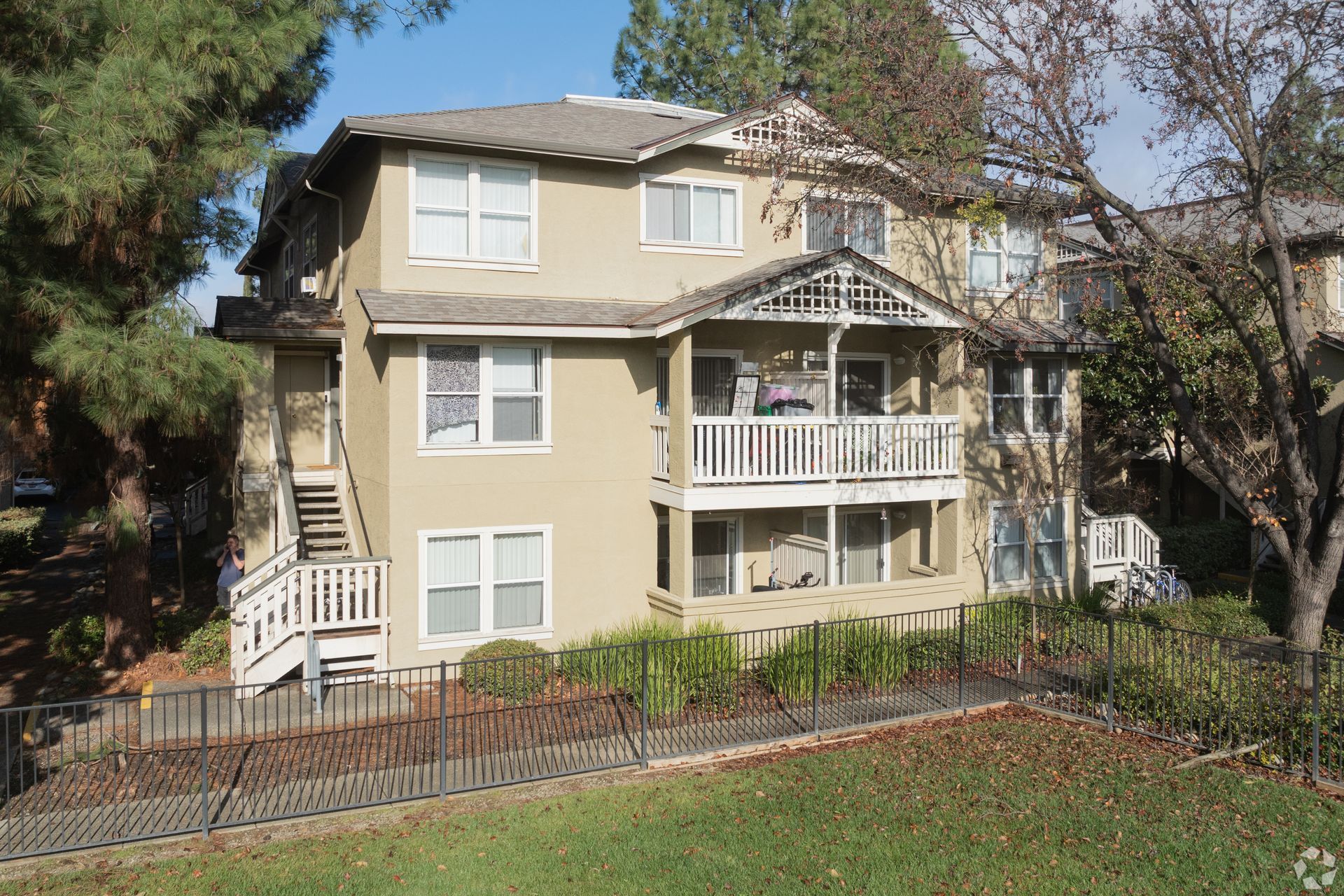 Beige two-story apartment building with white balconies and stairs. Surrounded by trees and a wrought iron fence.