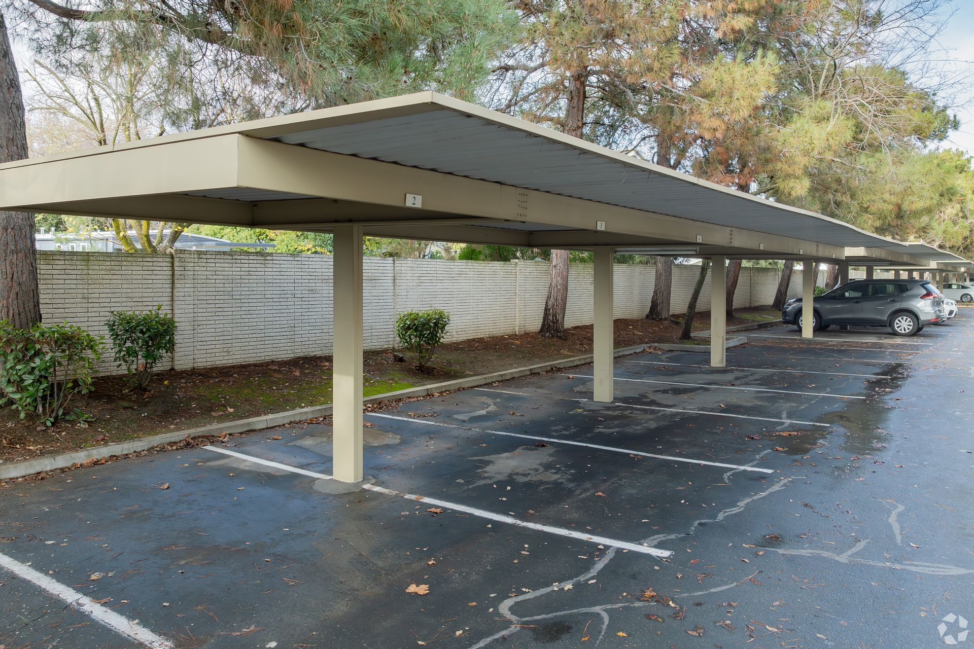 Covered parking area with concrete pillars, vehicles, and a low wall in the background.