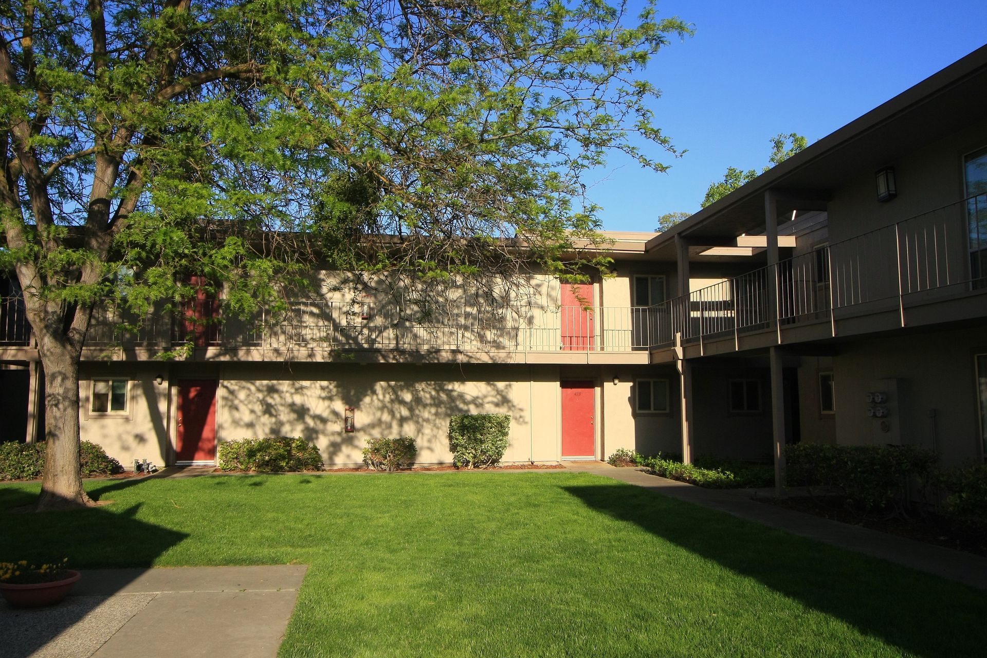 Apartment complex with a grassy courtyard, beige buildings, red doors, and a tree.