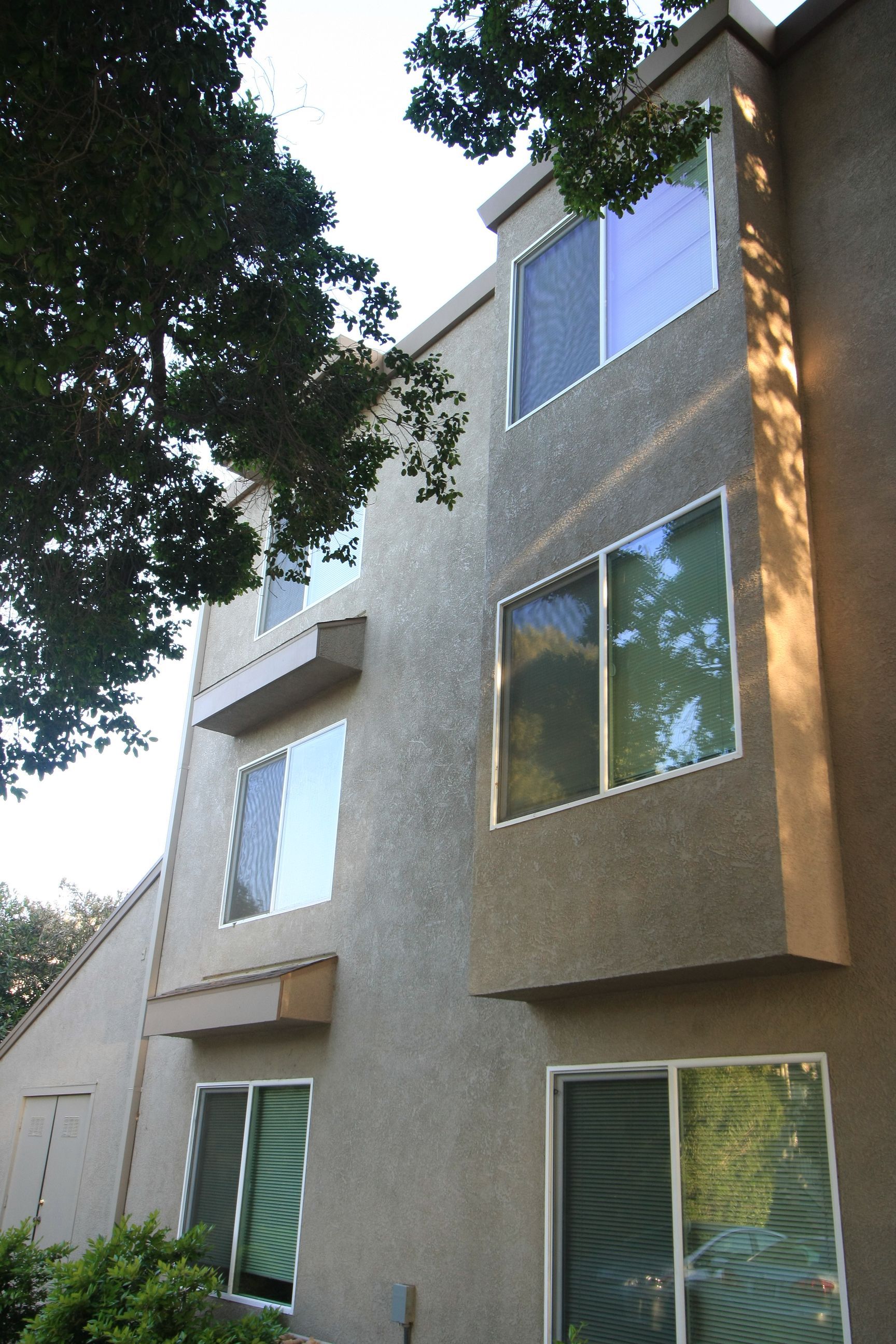 Three-story apartment building with stucco exterior and large windows; tree branches frame the left side.