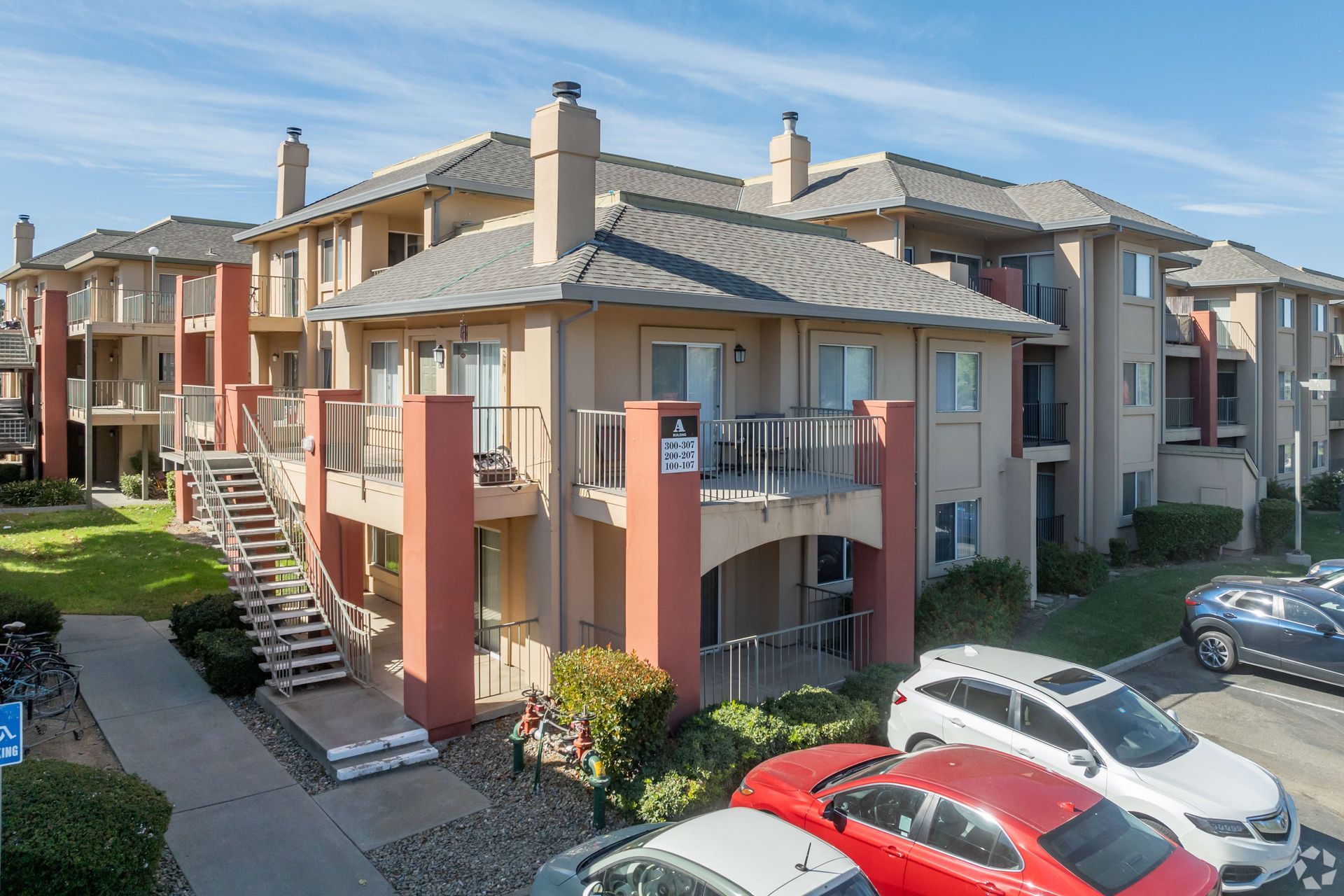 Apartment complex with multiple stories, balconies, stairs, and parked cars under a blue sky.