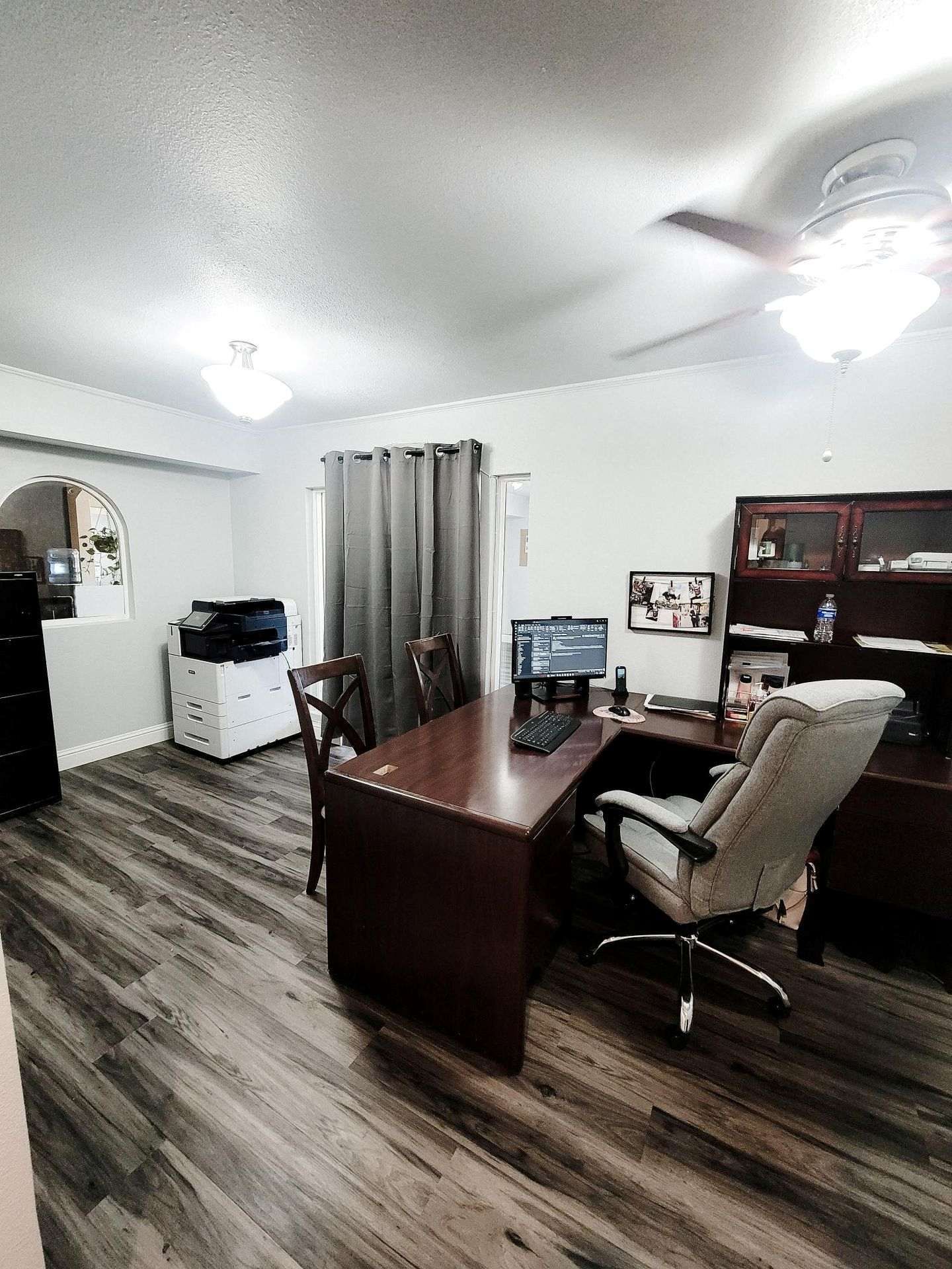Office interior with a desk, chair, printer, and filing cabinet. Light wood floors, white walls, and a ceiling fan.