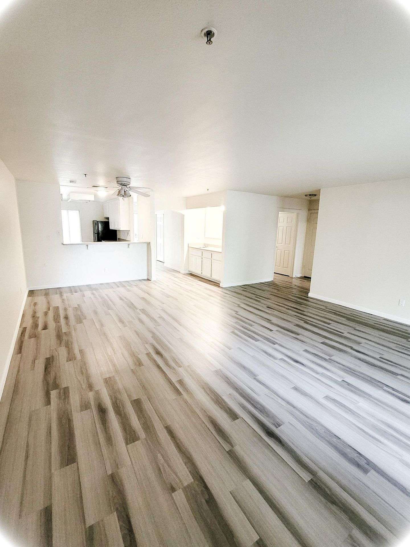 Open-concept living space with light wood-look flooring and white walls. Kitchen visible in the background.