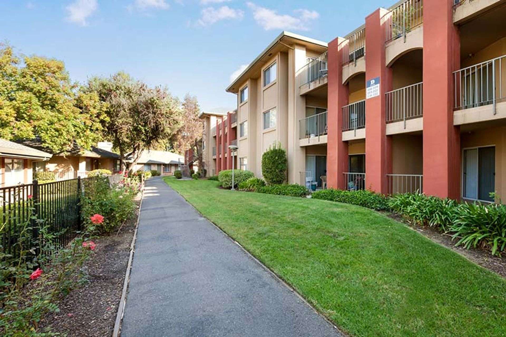 Apartment complex with a walkway, green grass, and trees under a blue sky.