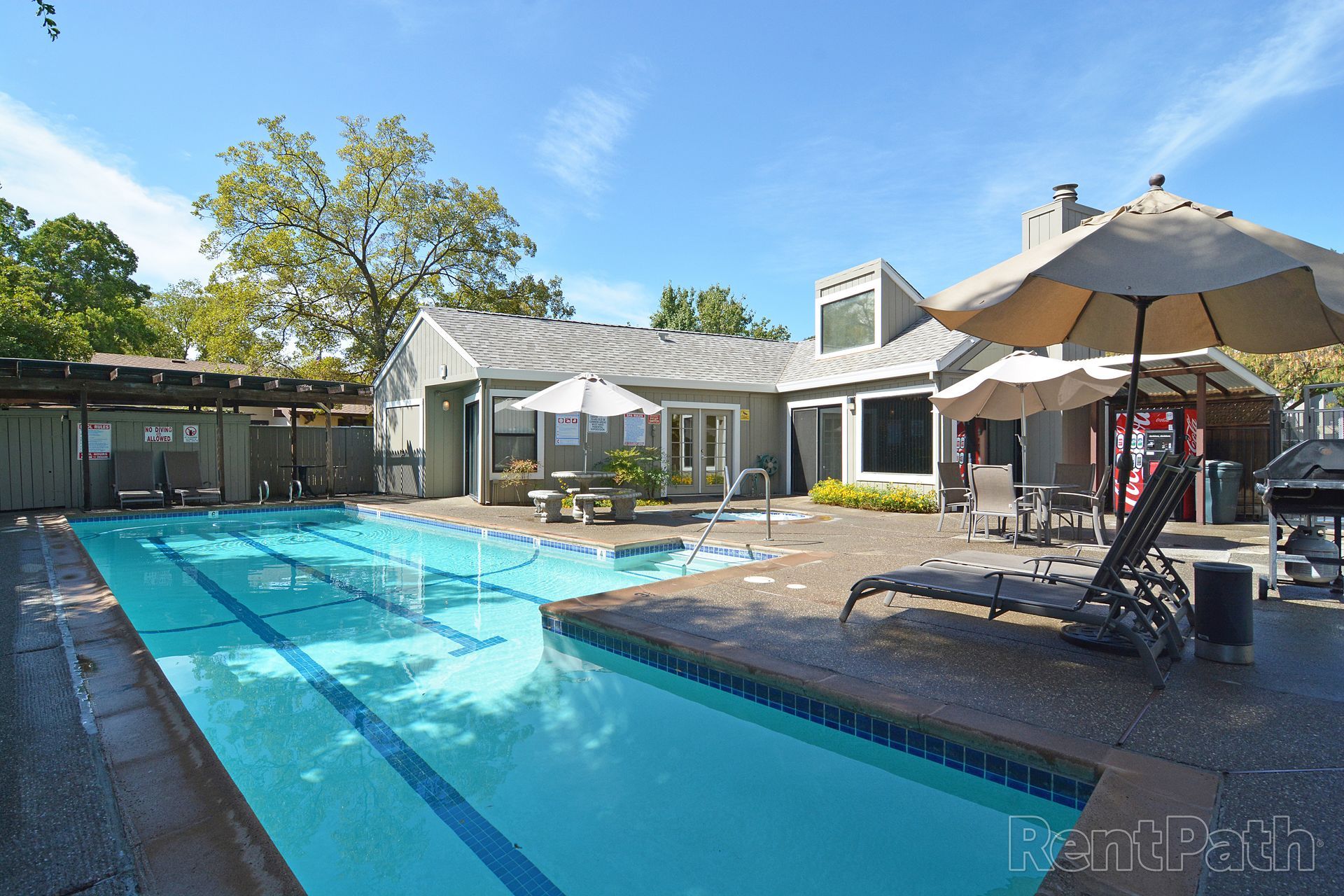 Outdoor swimming pool with a building, lounge chairs, and umbrellas under a blue sky.