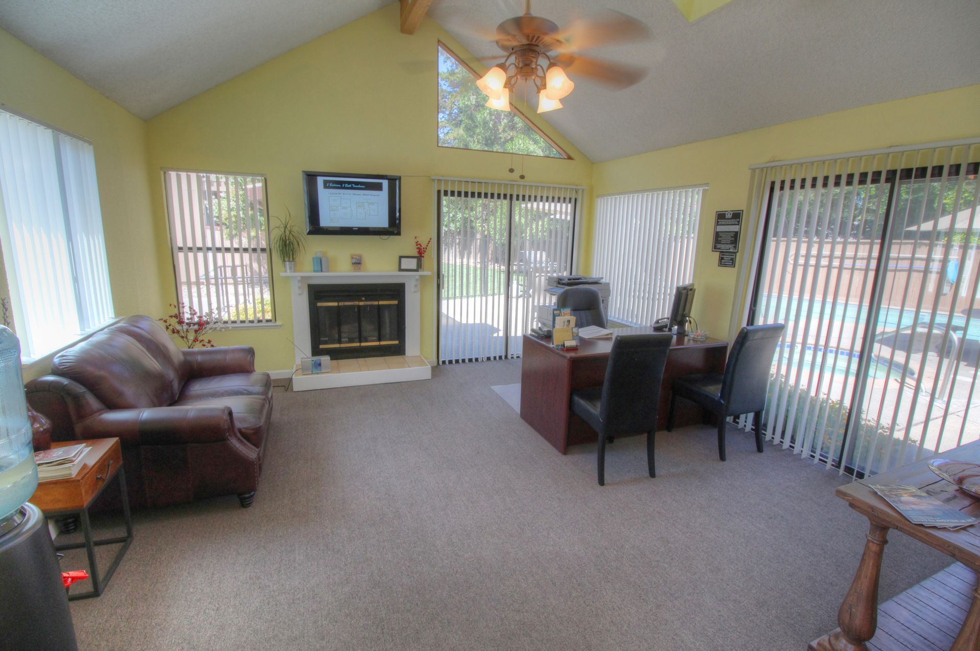 Living room with vaulted ceiling, fireplace, sliding glass door, brown leather sofa, and office setup.