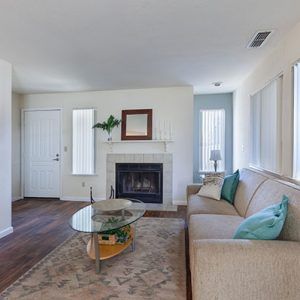 Living room with fireplace, beige sofa, glass coffee table, and area rug. White walls, wood floor.