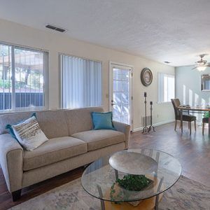 Living room with beige sofa, glass coffee table, and dining table, with sunlight streaming through windows.