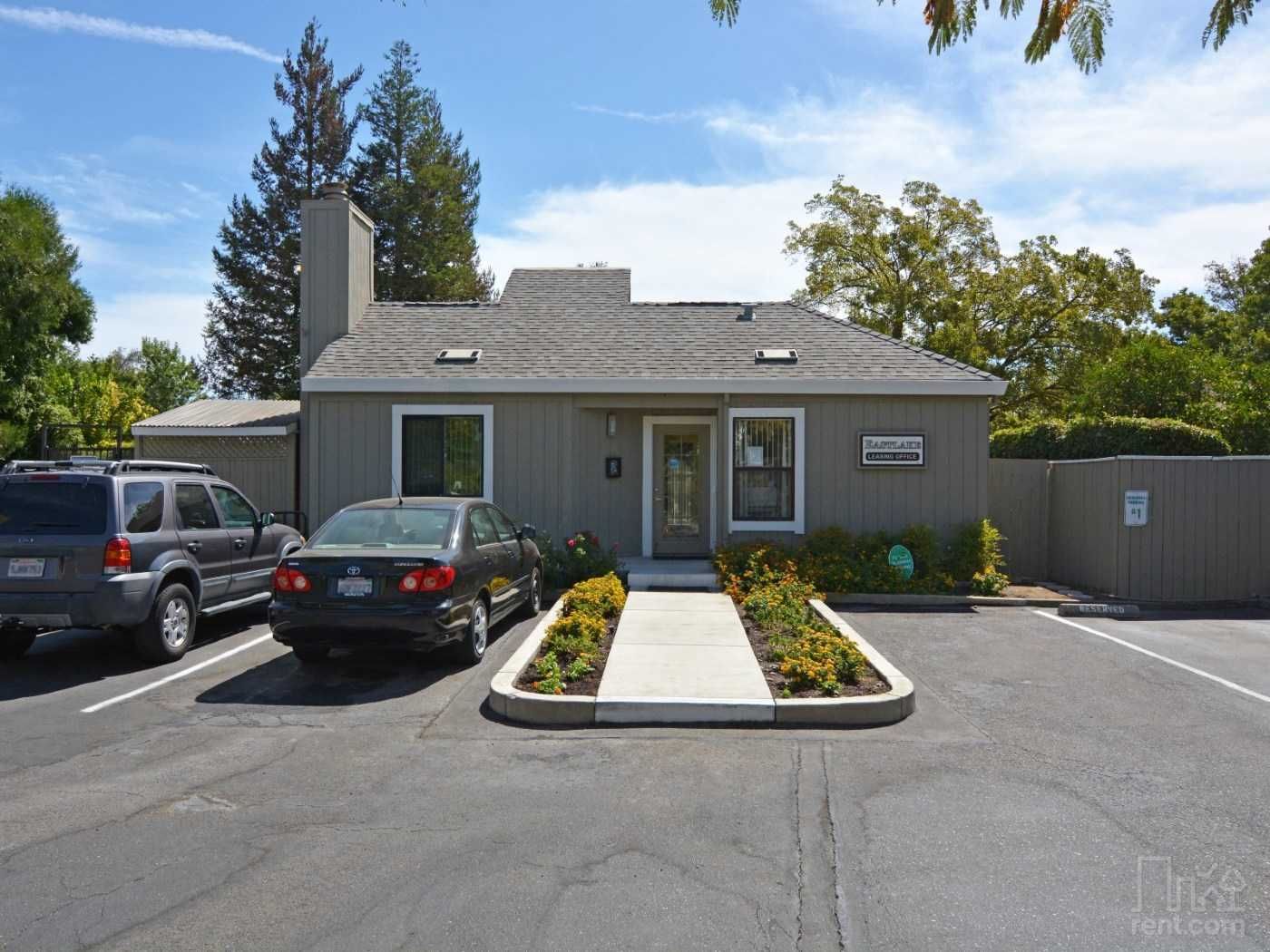 Gray building with cars parked in front; blue sky, trees.