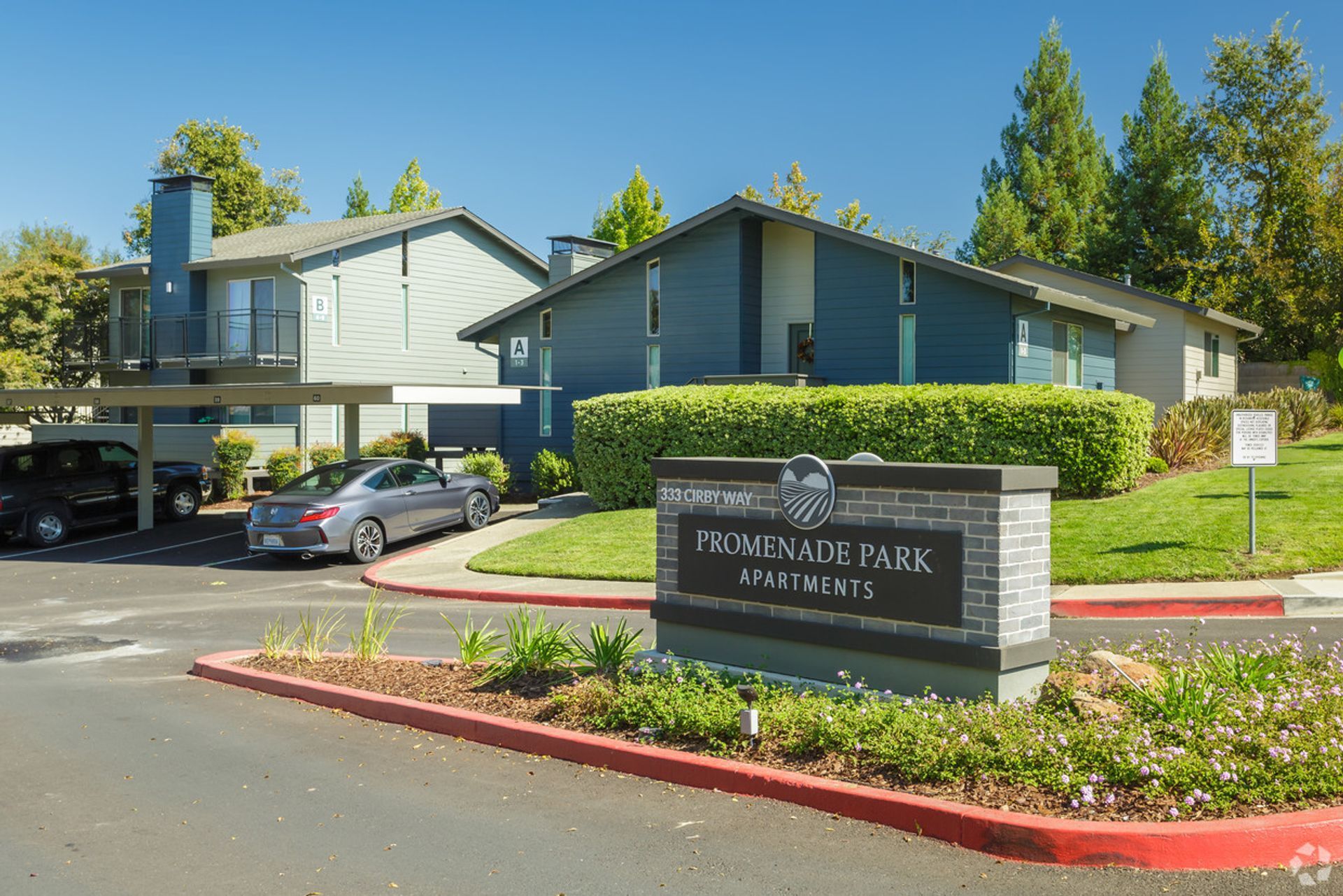 Apartment complex entrance sign with blue buildings, cars parked, and landscaping.