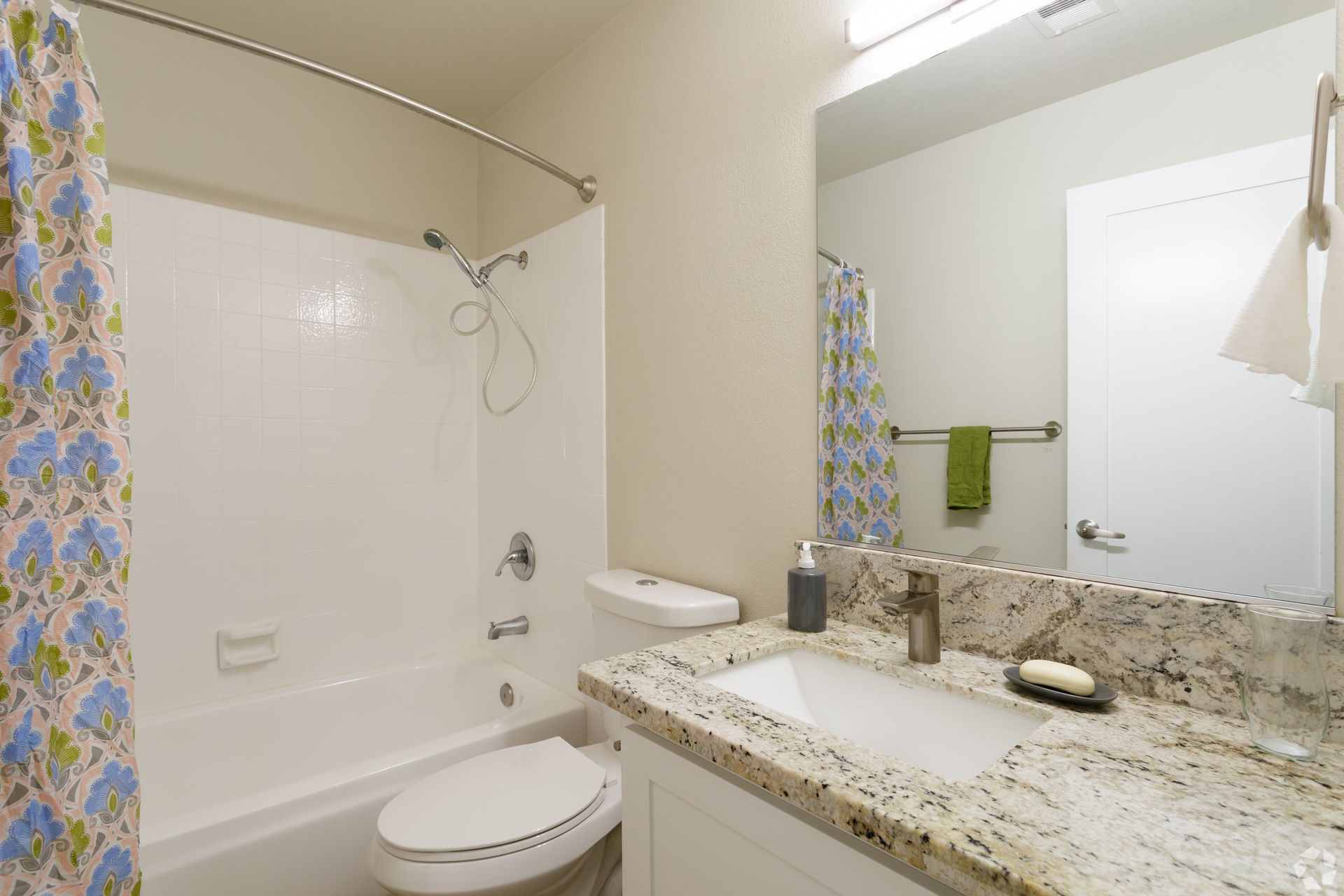 Bathroom with white tub, toilet, and vanity with granite countertop. Floral shower curtain.