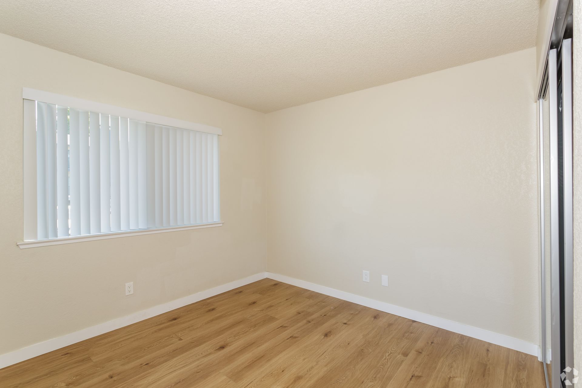 Empty room with light wood floors, window with blinds, and beige walls.