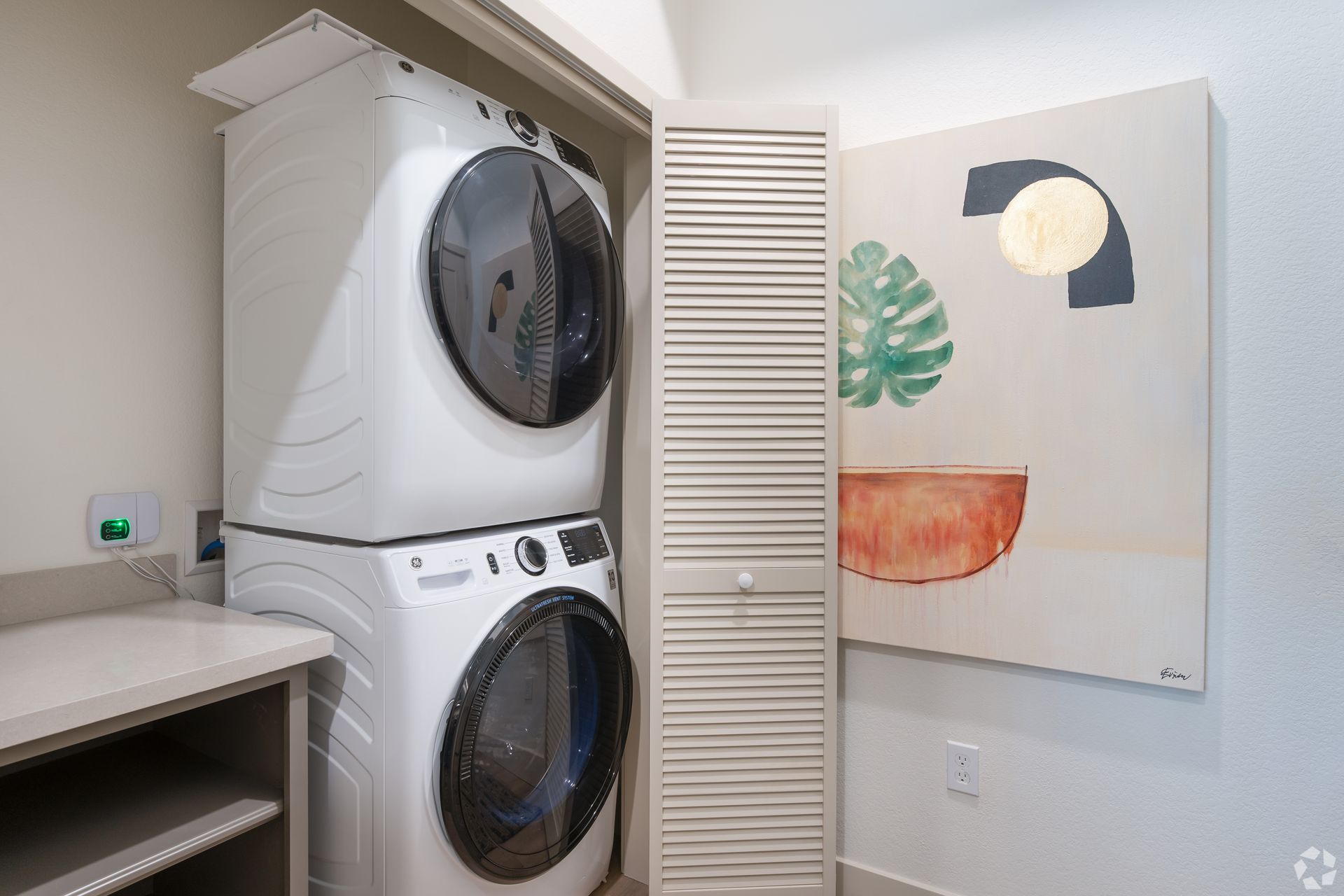 Stacked white washer and dryer in a closet with a louvered door and artwork on the wall.