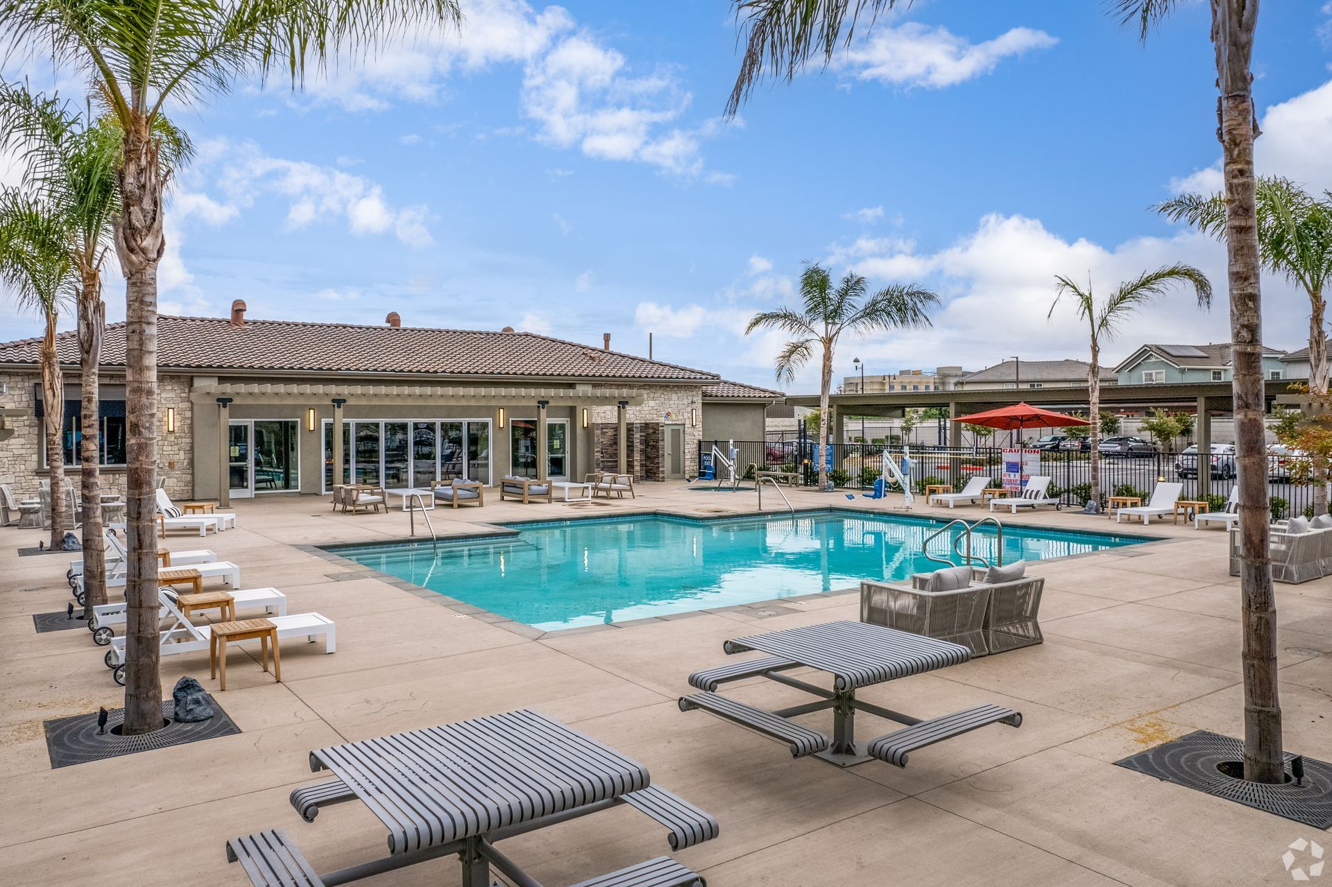 Pool area with a building, palm trees, and picnic tables under a blue sky.