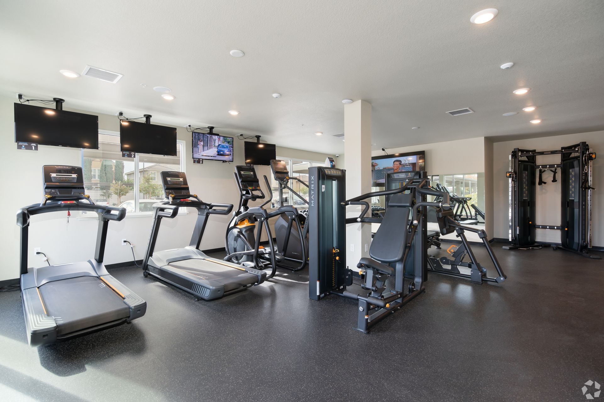 Gym with treadmills, elliptical, weight machines, and TVs. Black flooring, white walls, and bright lights.