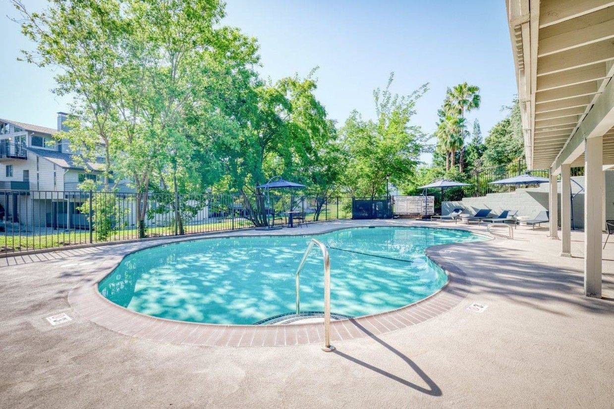 Pool with clear blue water surrounded by brick and a patio with chairs and a tree-filled background.