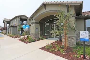 Luxe apartment complex entrance, tan building with stone accents, blue and white balloons, sunny day.