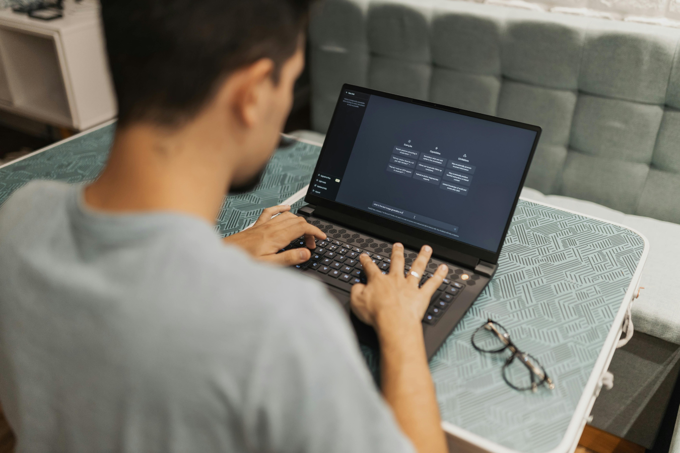 Man using a laptop at a desk, typing with both hands. Glasses rest nearby.