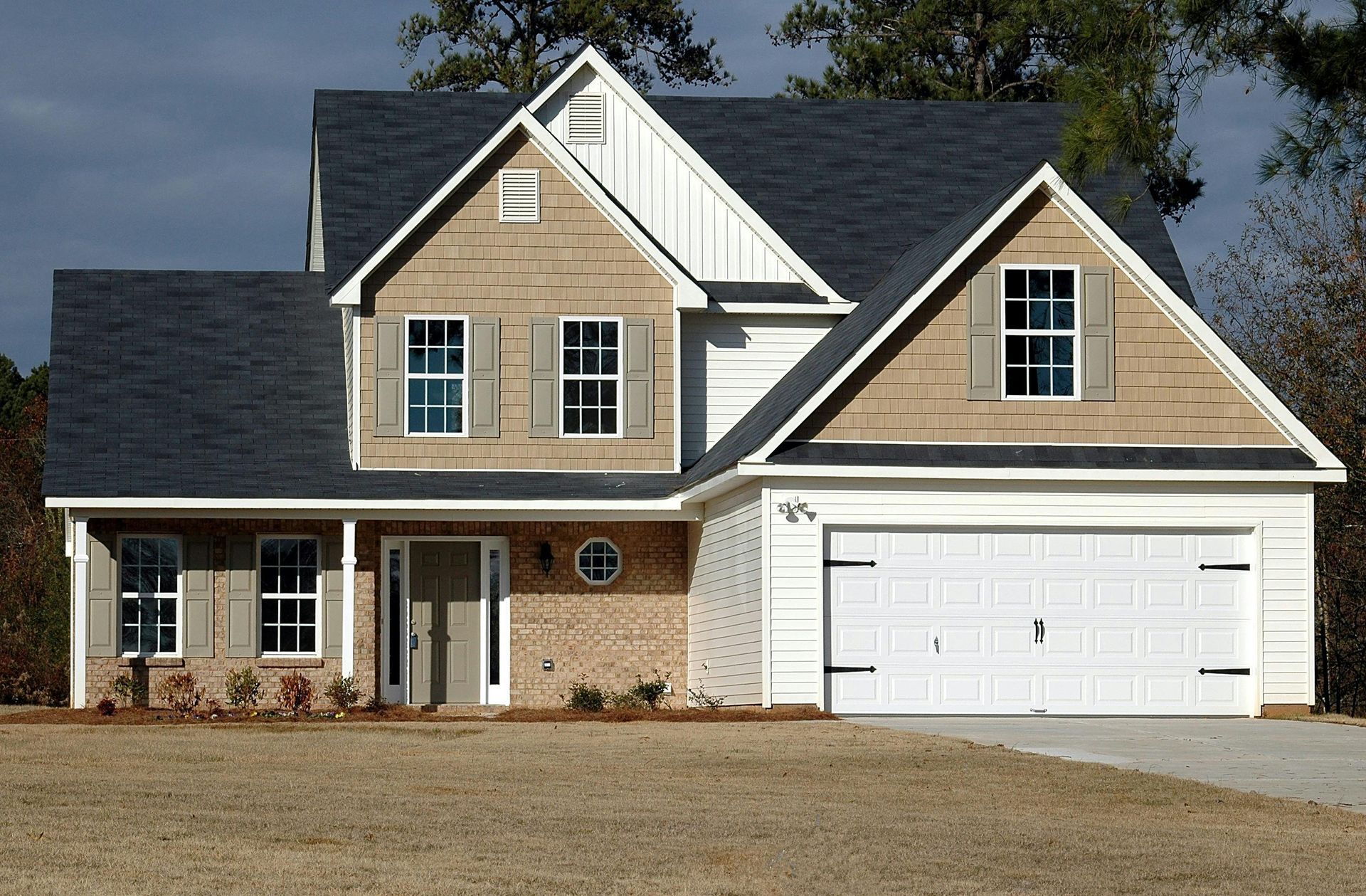 Two-story house with dark roof, tan siding, brick accents, and a white garage door, on a brown lawn.
