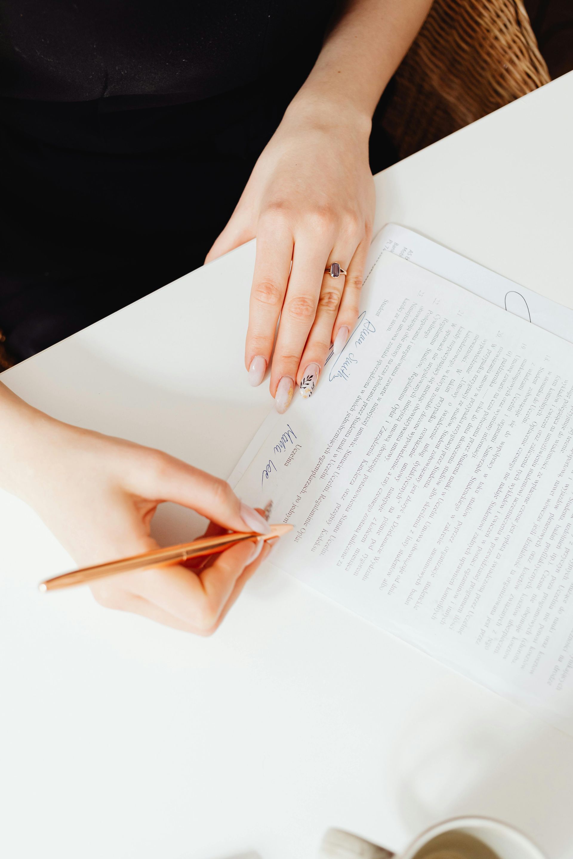 Person writing on a white paper with a pencil. One hand rests on the paper while the other writes, near a coffee cup.