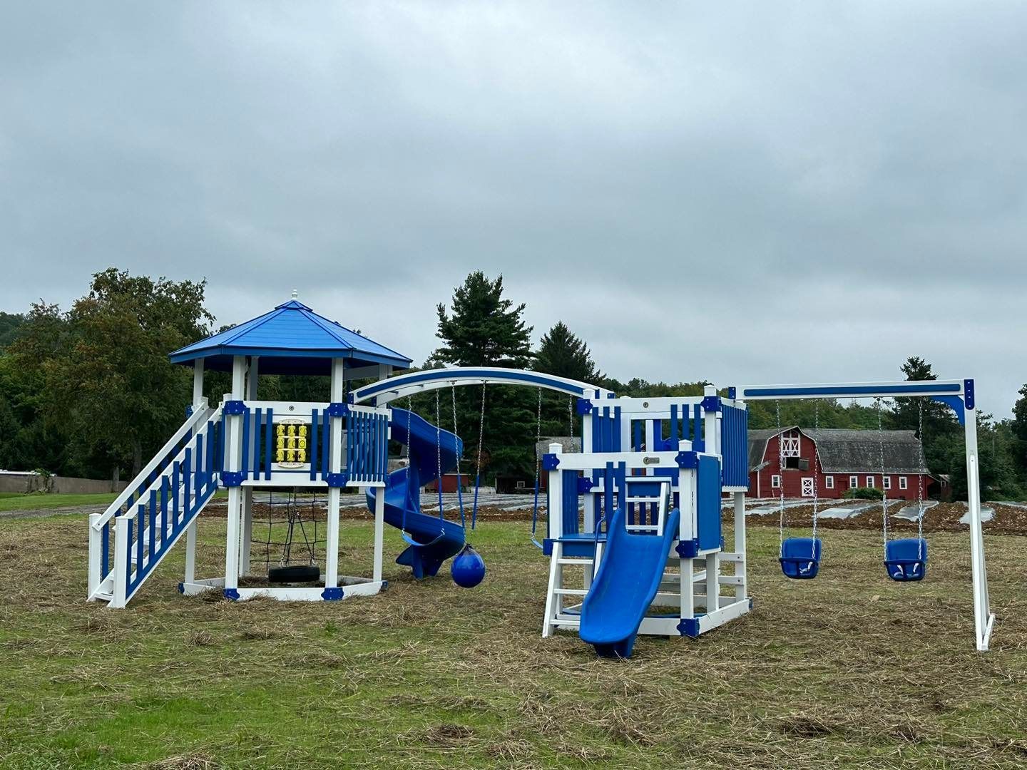 A blue and white playground set in a field with a red barn in the background.