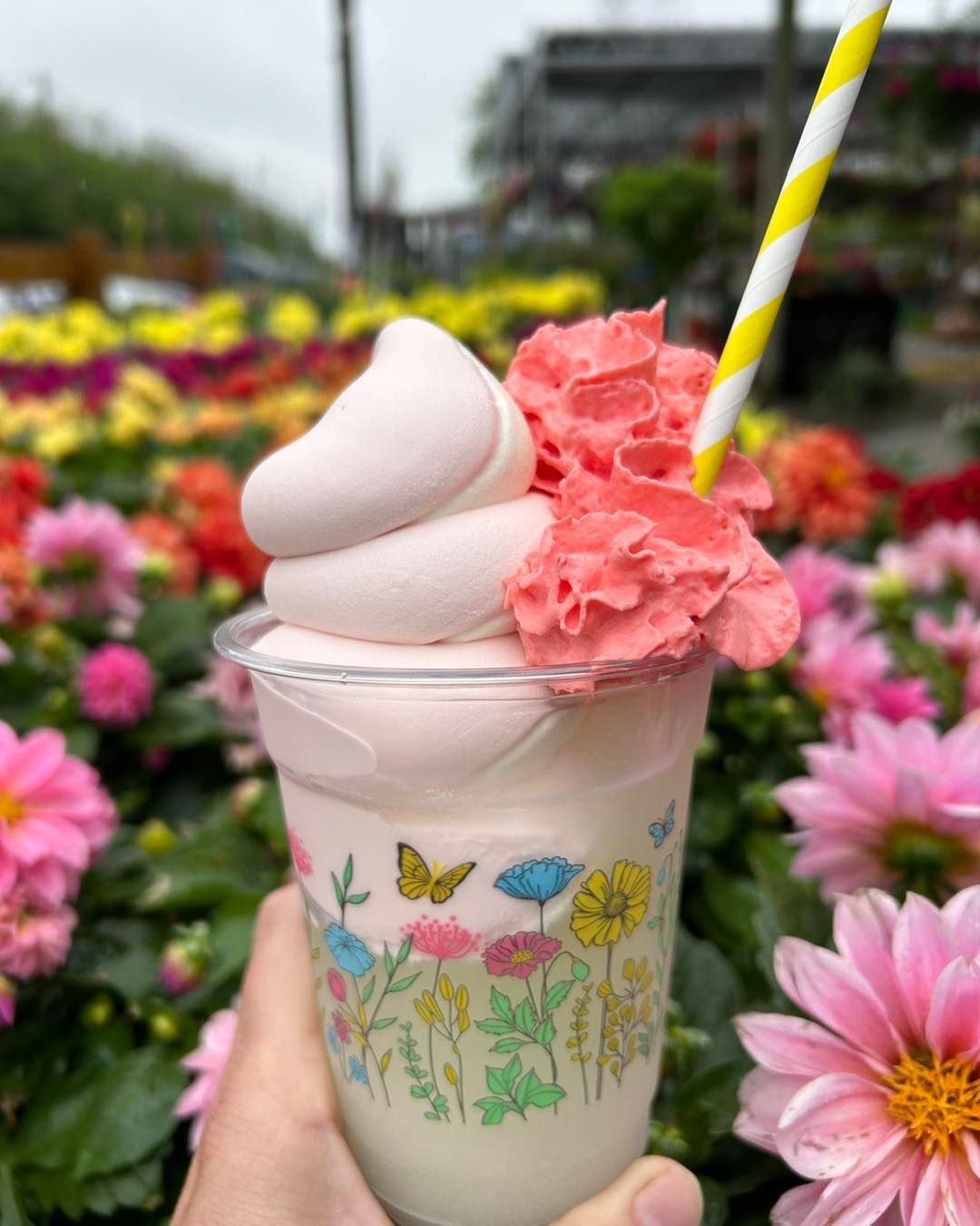 A person is holding a milkshake with a straw in front of flowers.