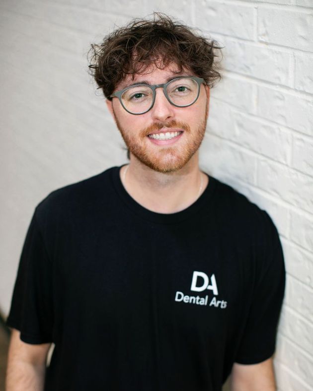 A man wearing glasses and a black shirt is standing in front of a white brick wall.