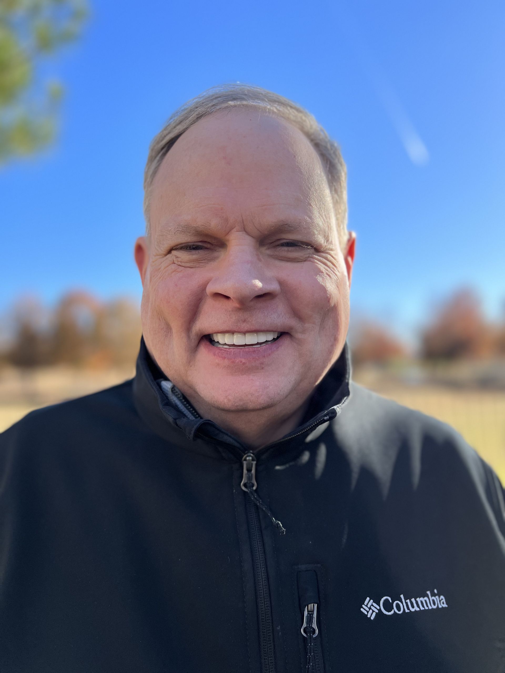 A man wearing a black columbia jacket is smiling for the camera.