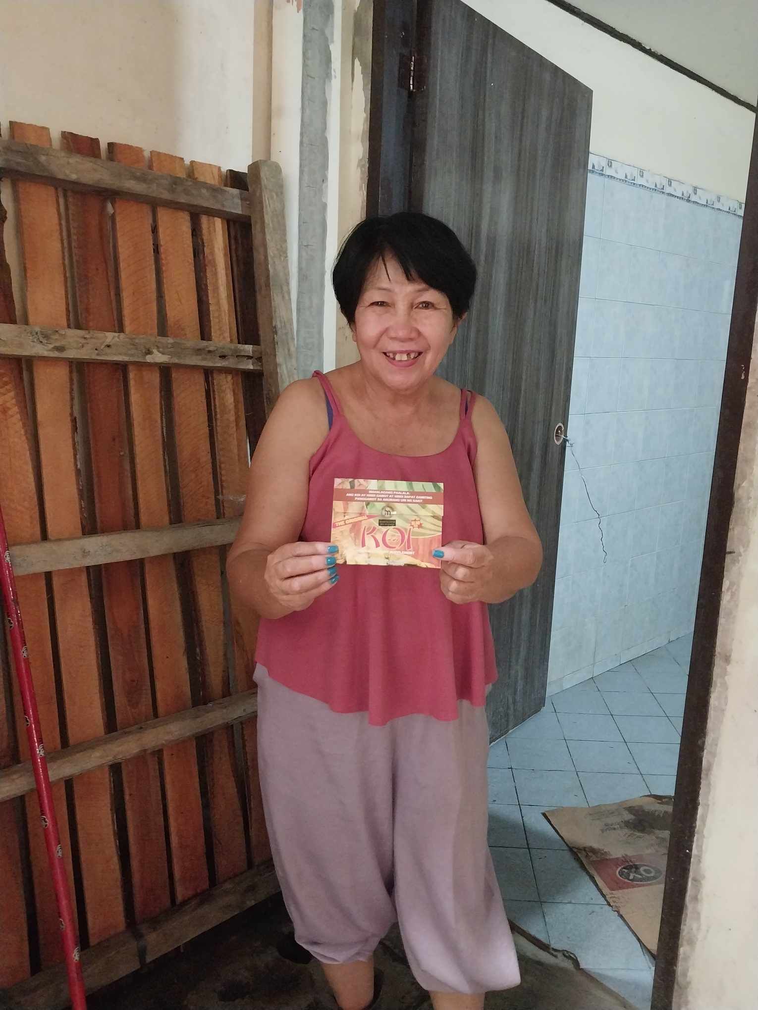 A woman in a pink tank top is holding a card in front of a wooden fence.