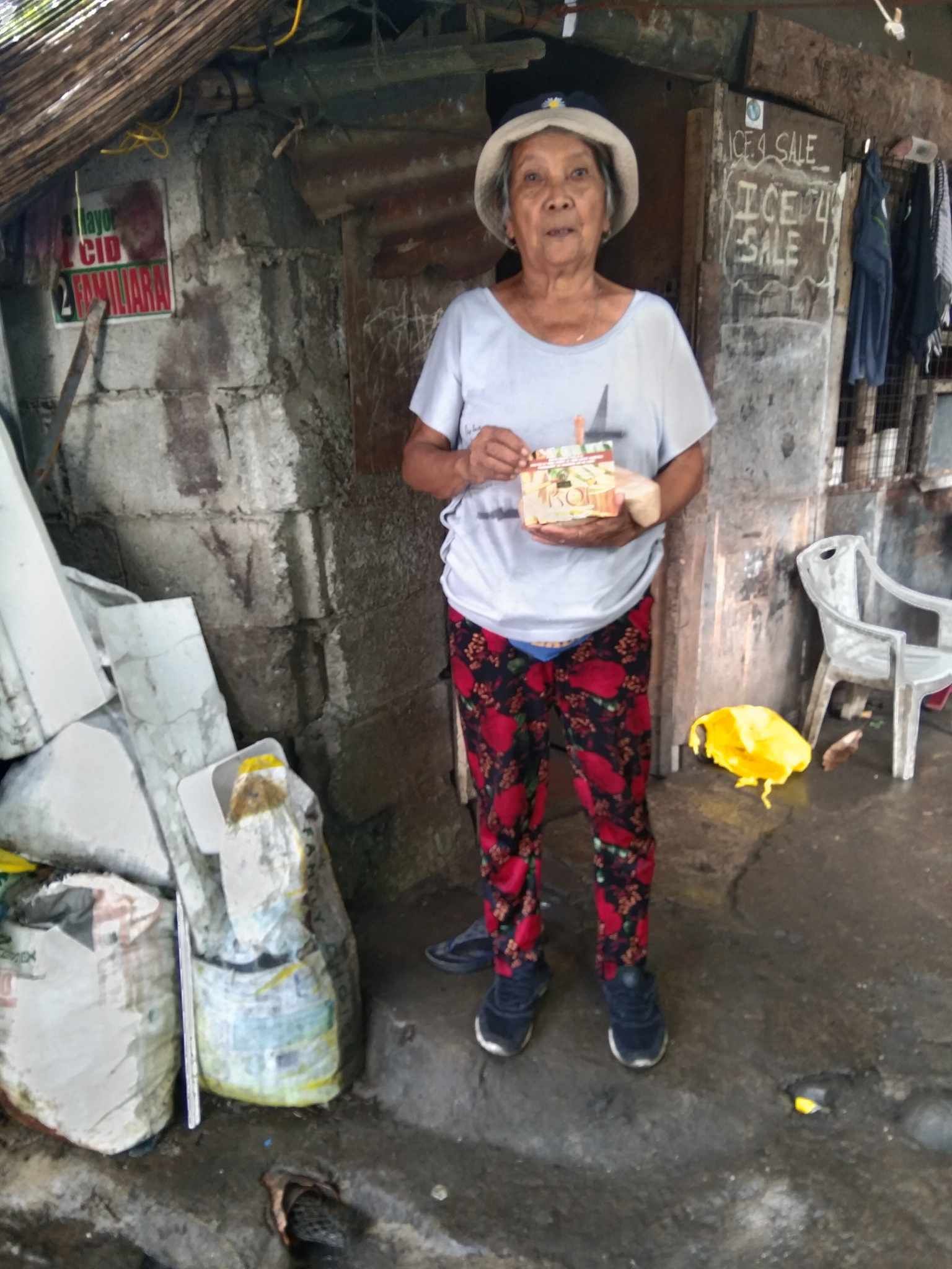 An elderly woman is standing in front of a building holding a cake.