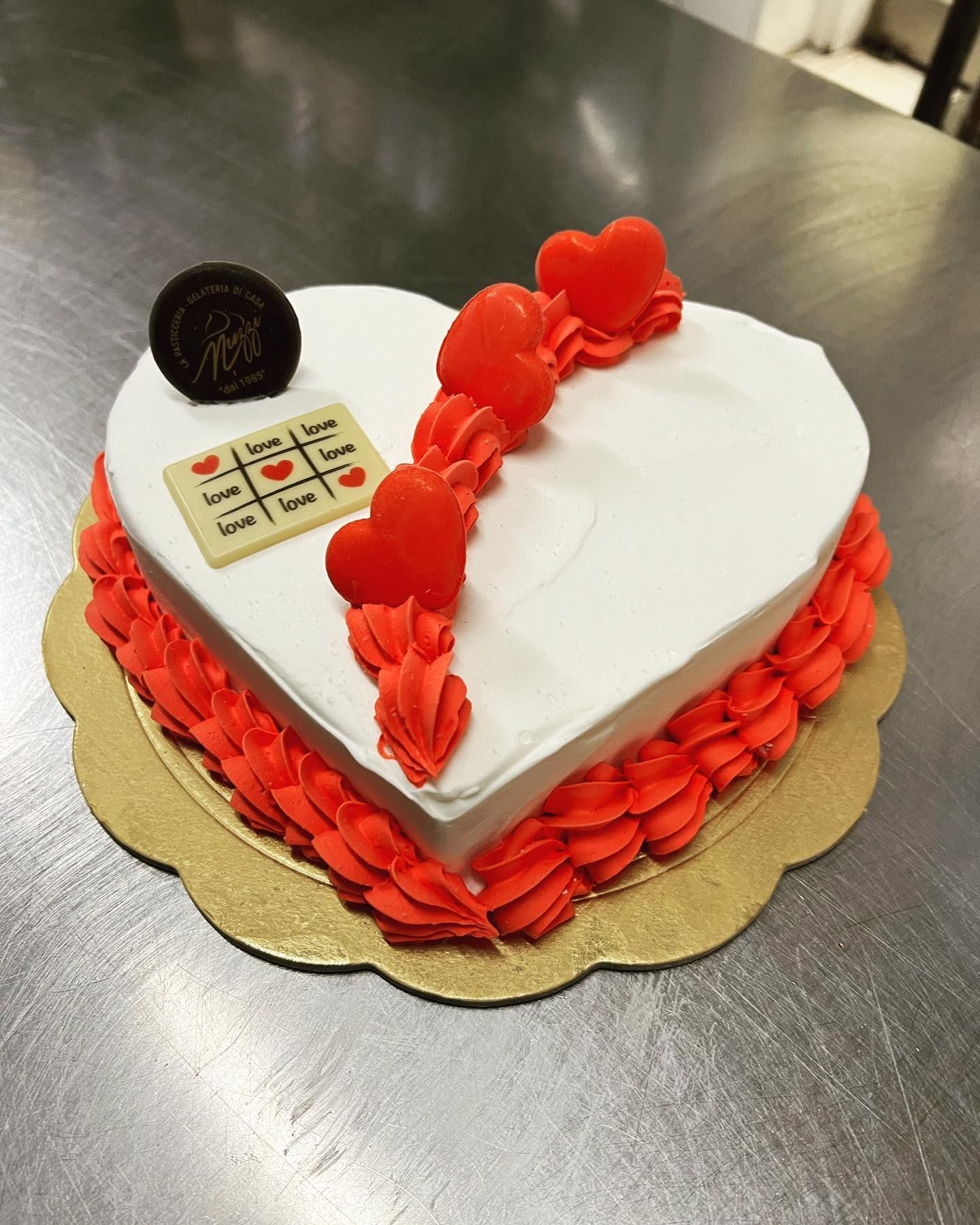 Torta a forma di cuore con glassa bianca, decorazioni rosse con sac à poche, cuoricini rossi e una placca di cioccolato con il gioco del tris su una base dorata.