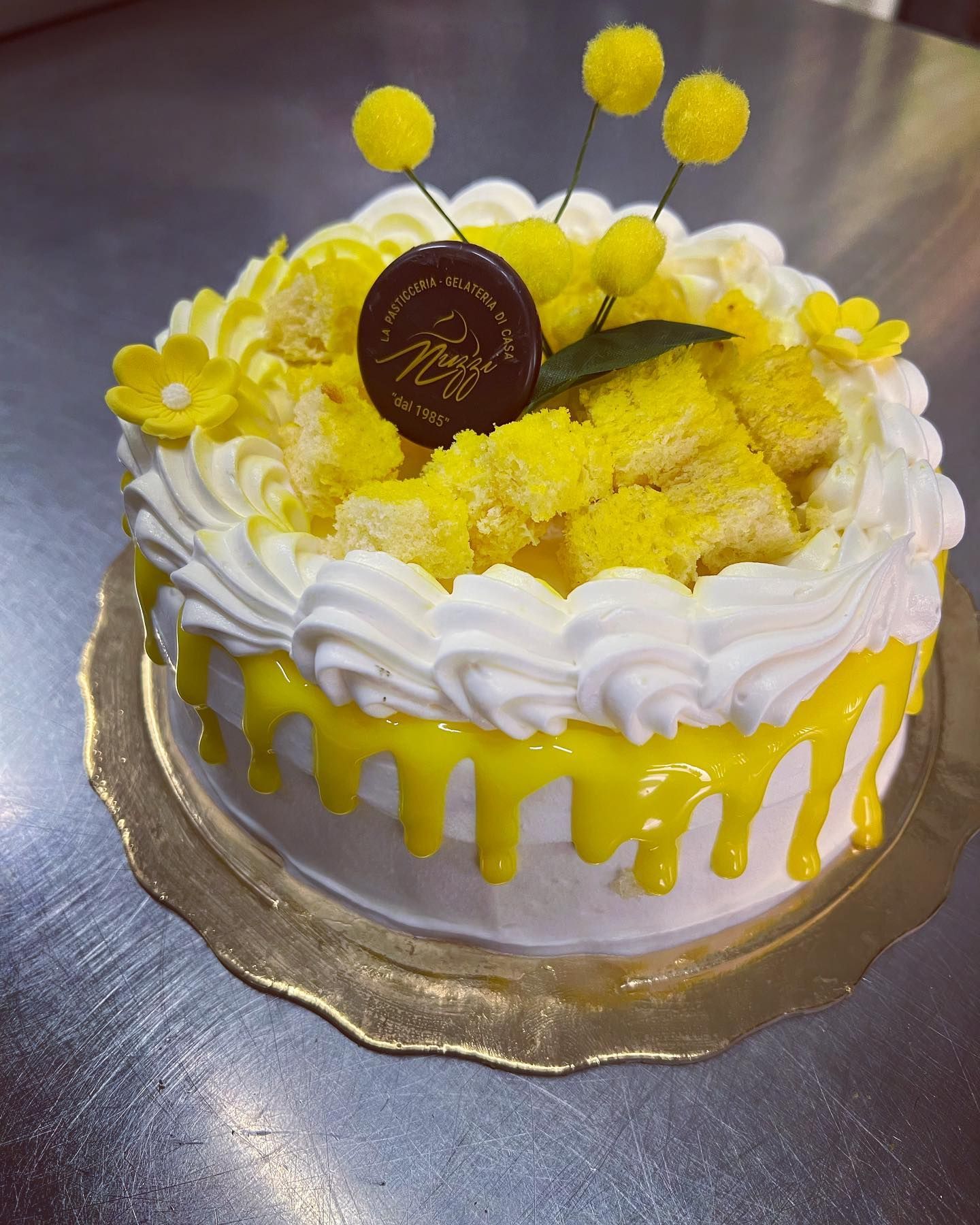 Una torta ricoperta di glassa bianca con colature di glassa gialla, decorata con briciole di torta, fiori gialli e un disco con il logo del marchio.
