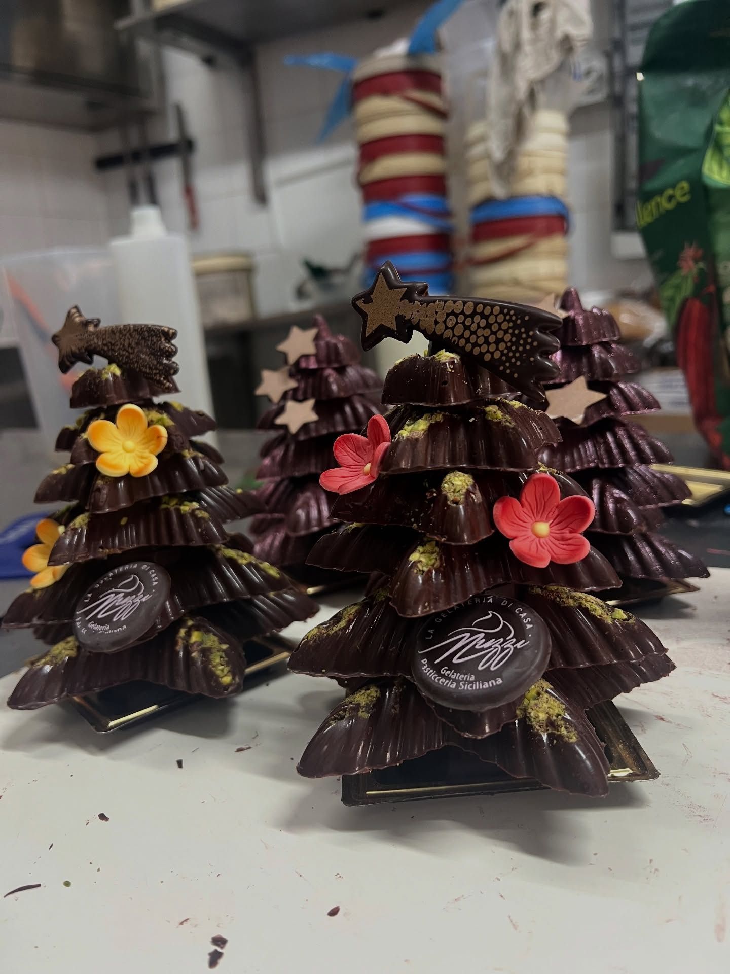 Tre alberi di Natale di cioccolato decorati con fiori commestibili e puntali a forma di stella, appoggiati su un bancone della cucina.