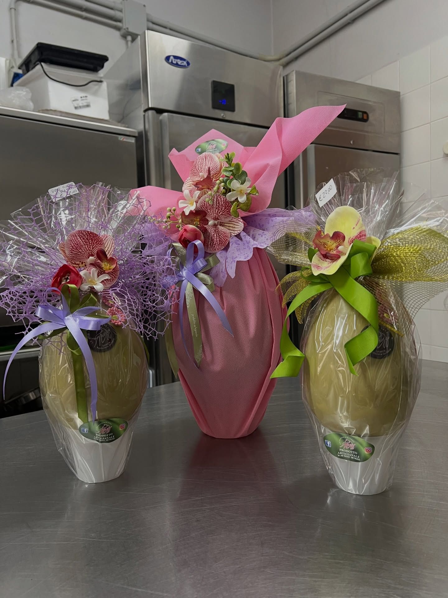 In una cucina professionale, tre uova di Pasqua decorate e avvolte in cellophane con motivi floreali sono appoggiate su una superficie metallica.