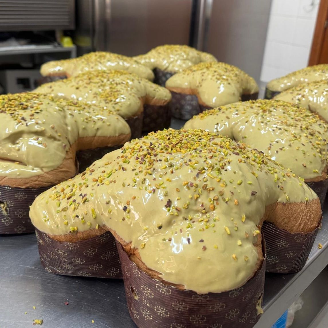 Colomba di Pasqua in stampini di carta, guarnite con crema al pistacchio e pistacchi tritati, disposte su un tavolo di metallo.